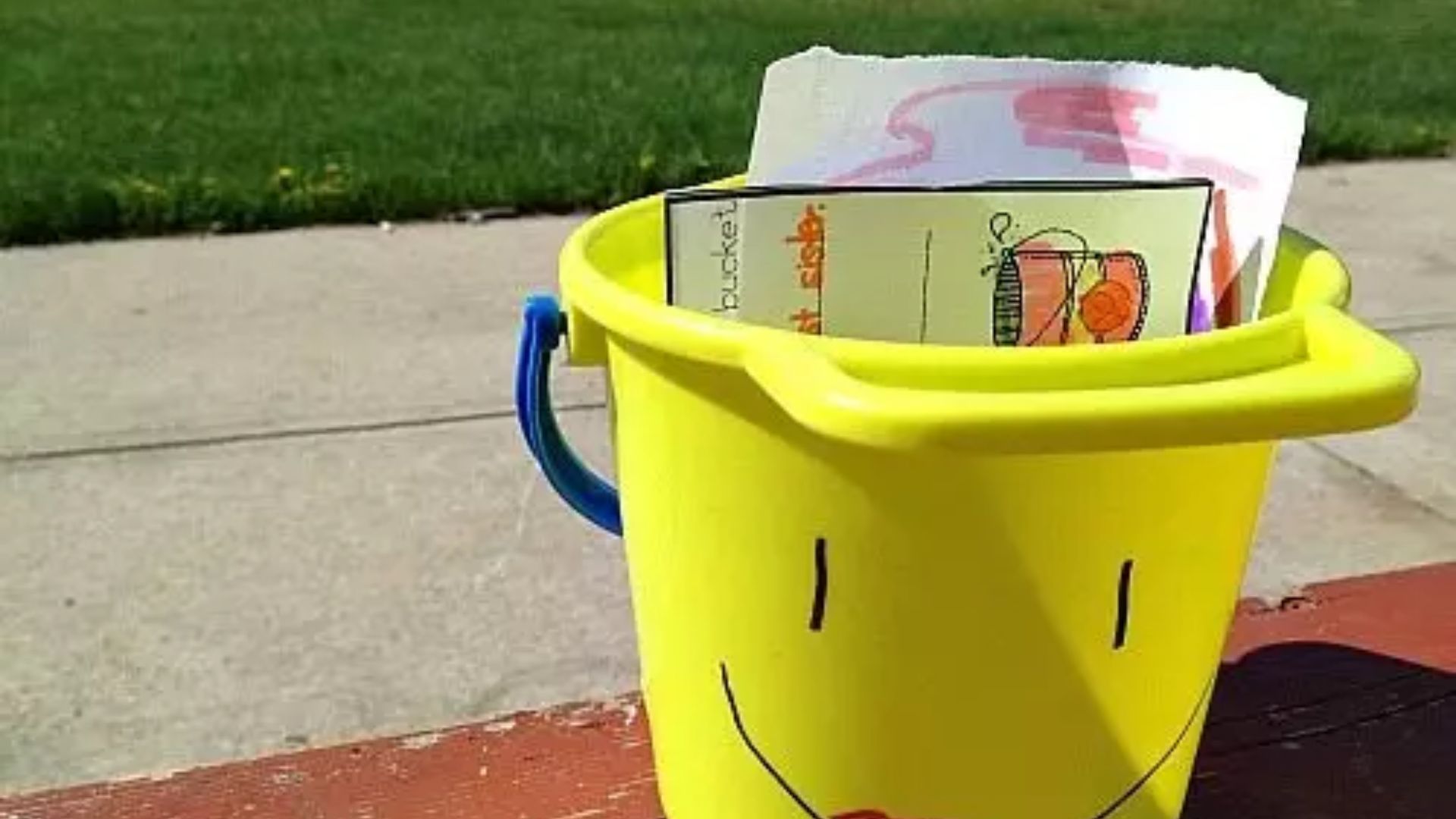 Yellow plastic bucket with a hand-drawn smiley face sitting on a red picnic table, filled with student drawings and bucket filler notes.