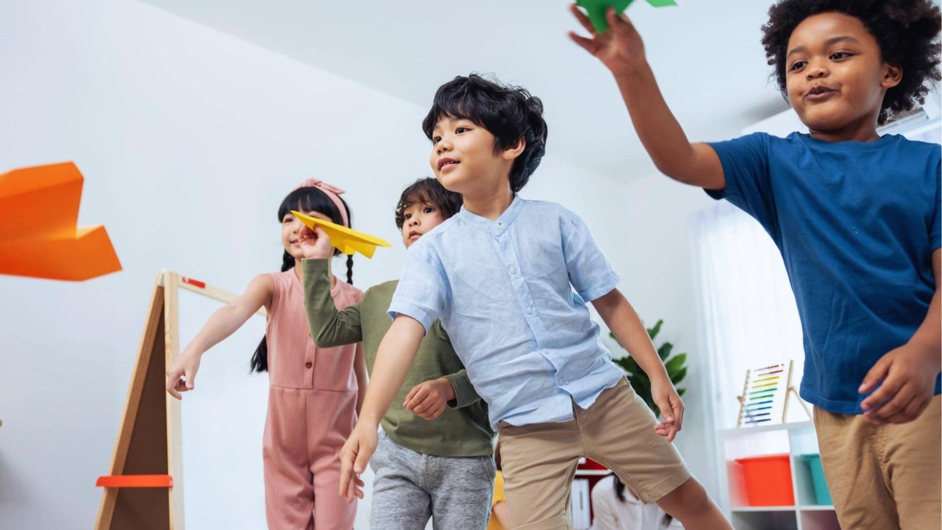 Group of kids indoors throwing paper airplanes, smiling, white room, easel, playful.