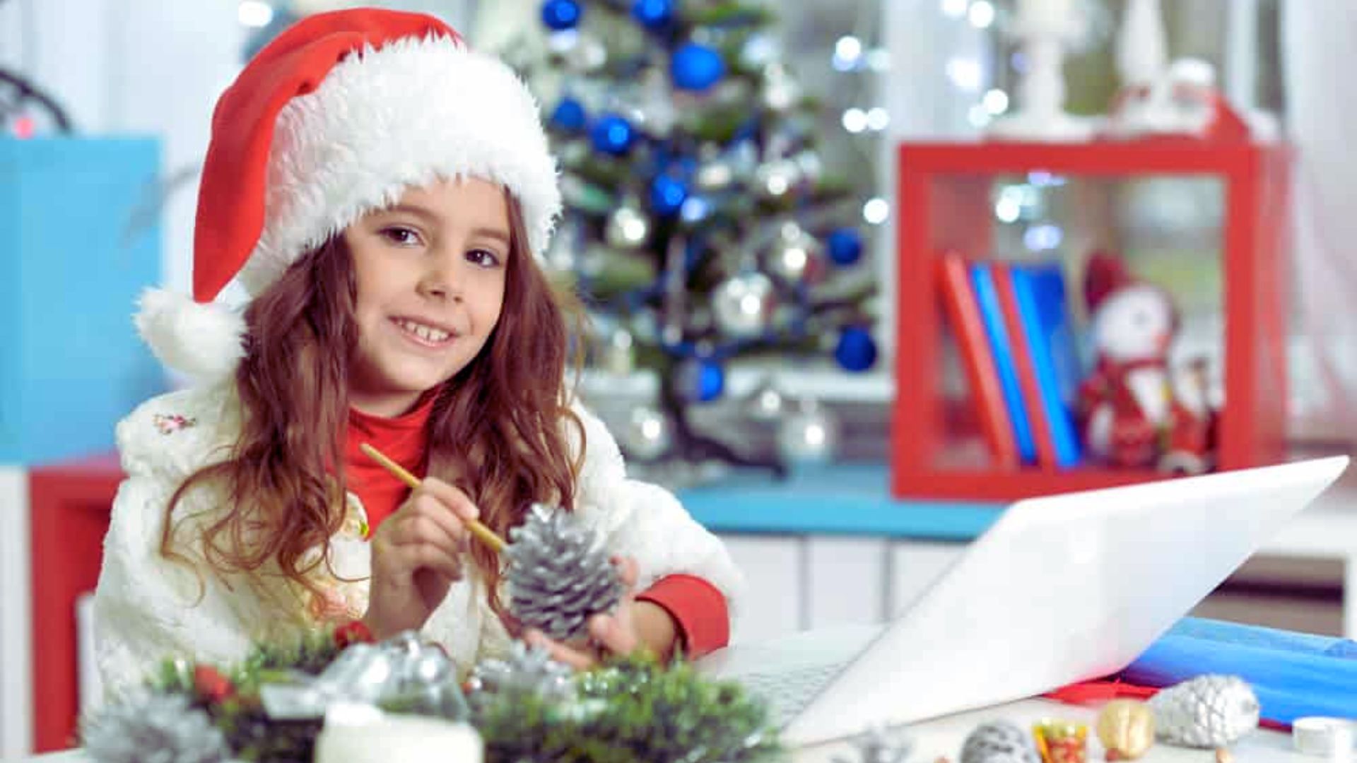 A young girl wearing a Santa hat is focused on using a laptop at a festive setting.