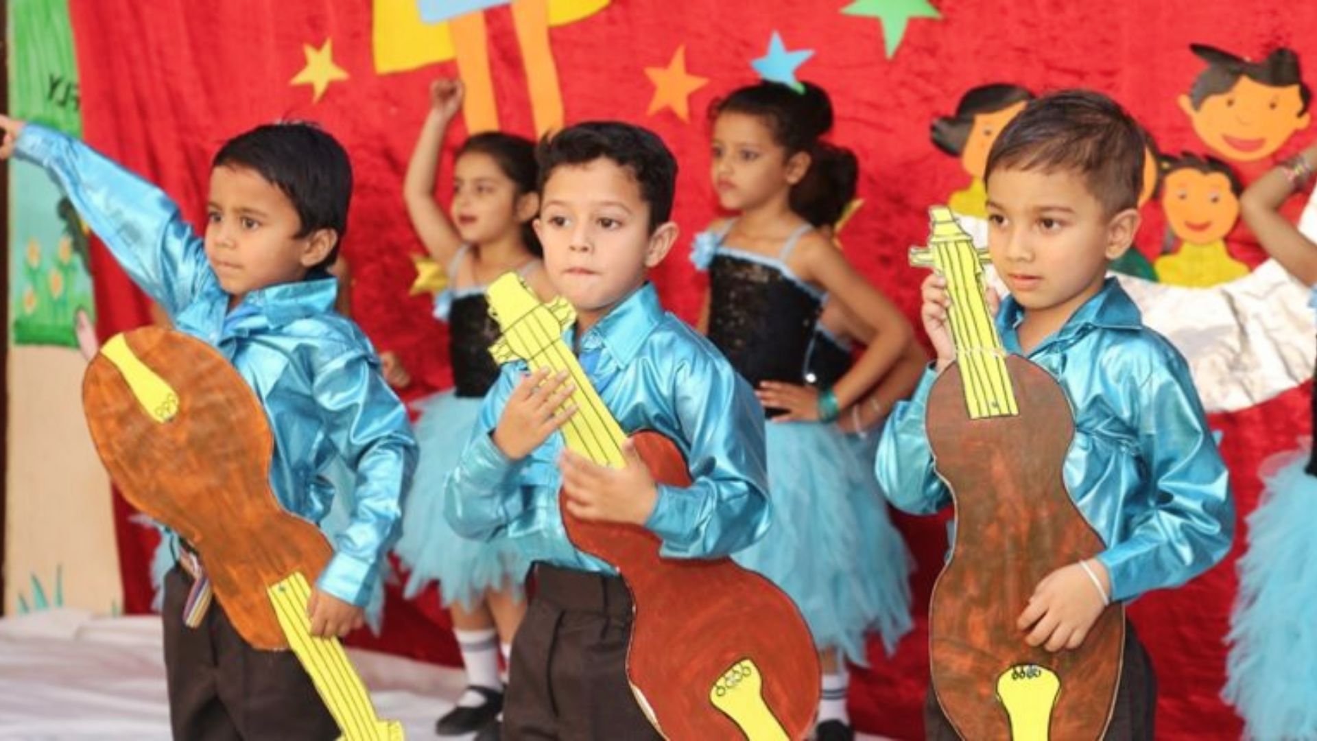 A group of children dressed in blue and black outfits, each holding a guitar, participating in cultural activities.