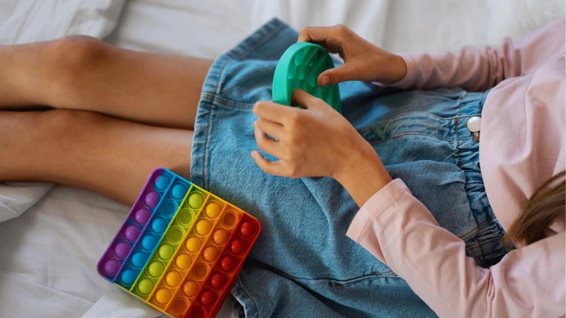 Girl in pink shirt presses green pop it fidget toy, rainbow one nearby, Activities for Autistic Kids.
