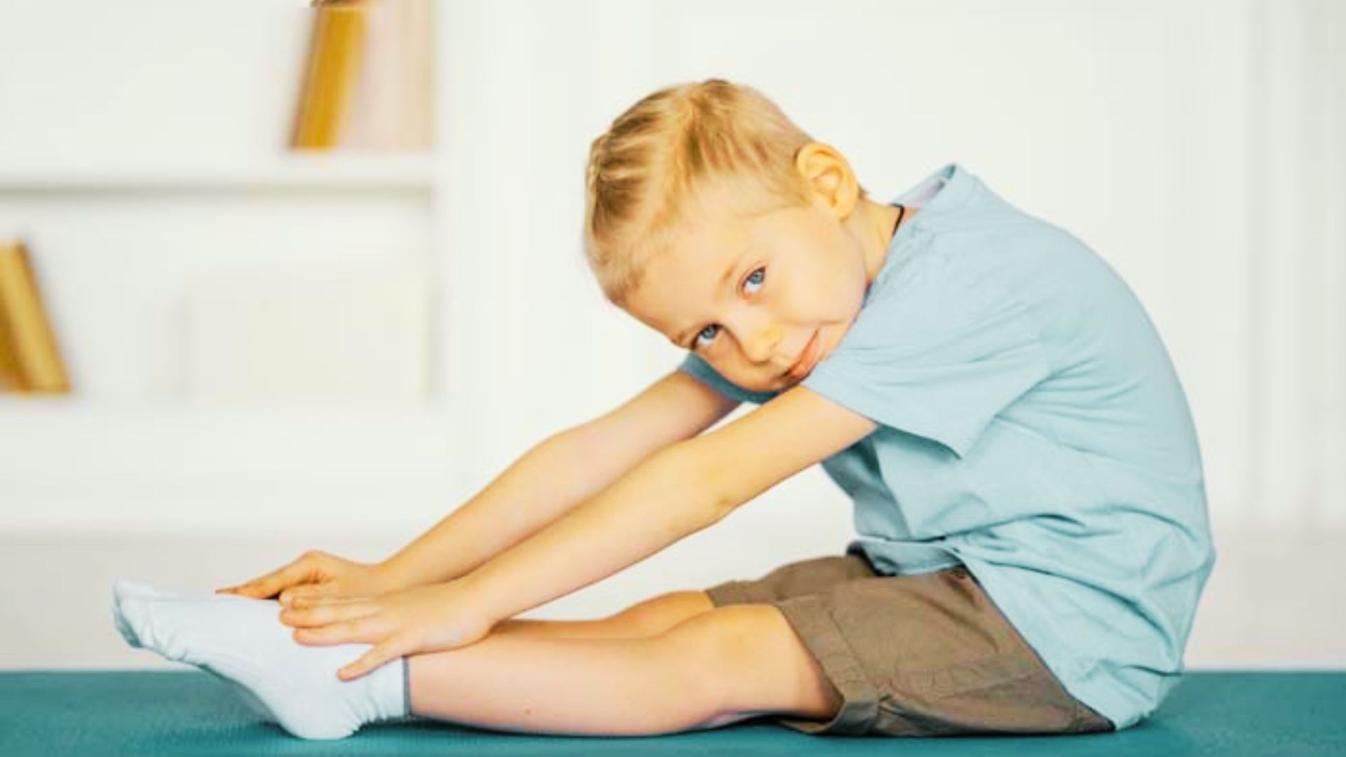 A young boy sits on a blue mat touching his toes looking content and relaxed in a bright, open space.