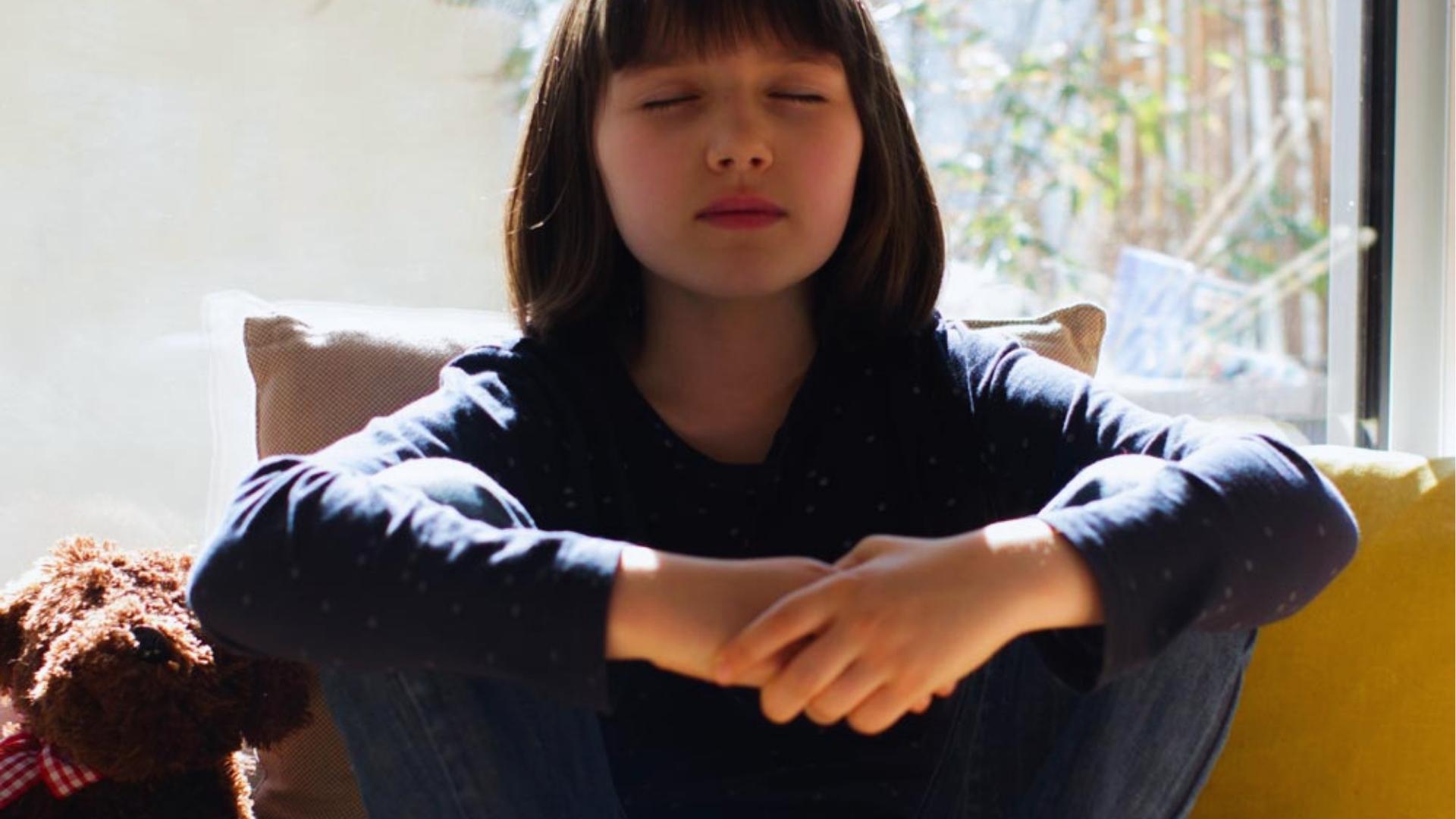 Girl sits cross legged with eyes closed meditating on couch, teddy nearby, sunlight, Activities for Autistic Kids.