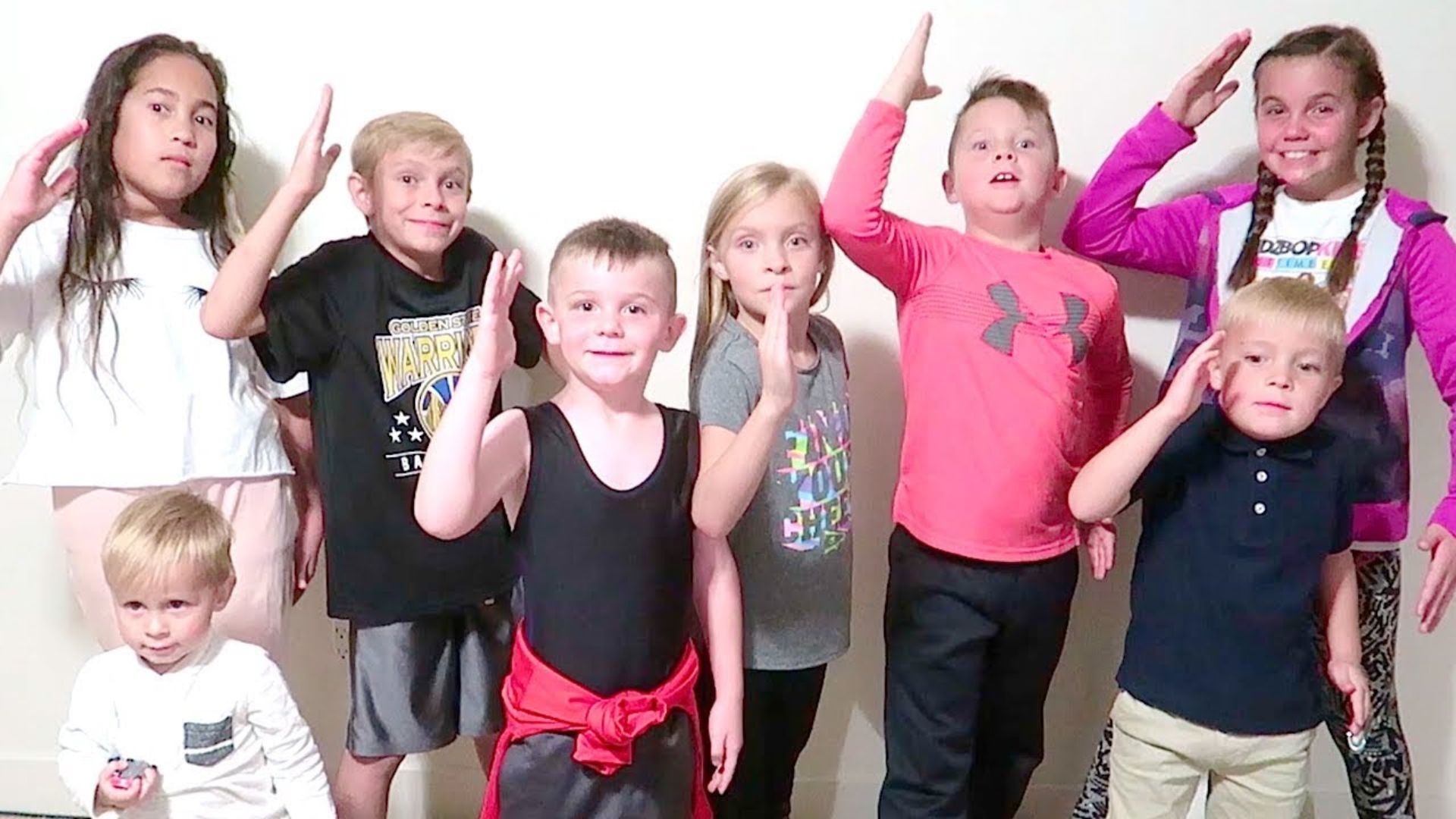 Eight kids of different ages standing against a wall making fun hand gestures together, playing a group movement game for kids at home