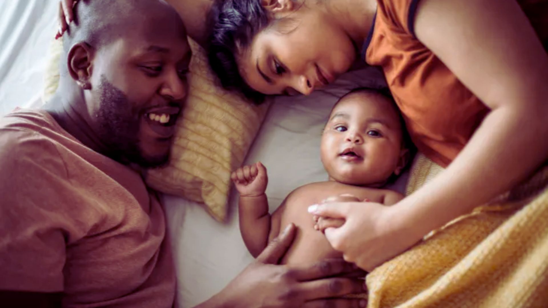 A man and woman lie on a bed, gently cradling a sleeping baby between them.