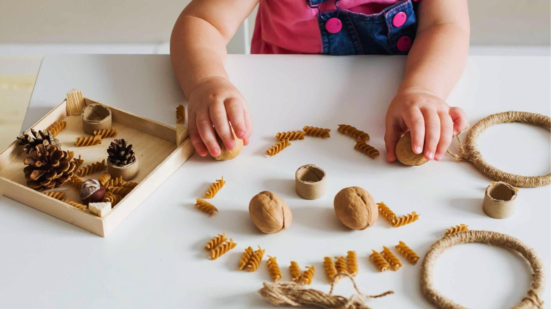 Toddler arranges walnuts and pasta pieces on white table for sensory craft, natural materials, Activities for Autistic Kids.