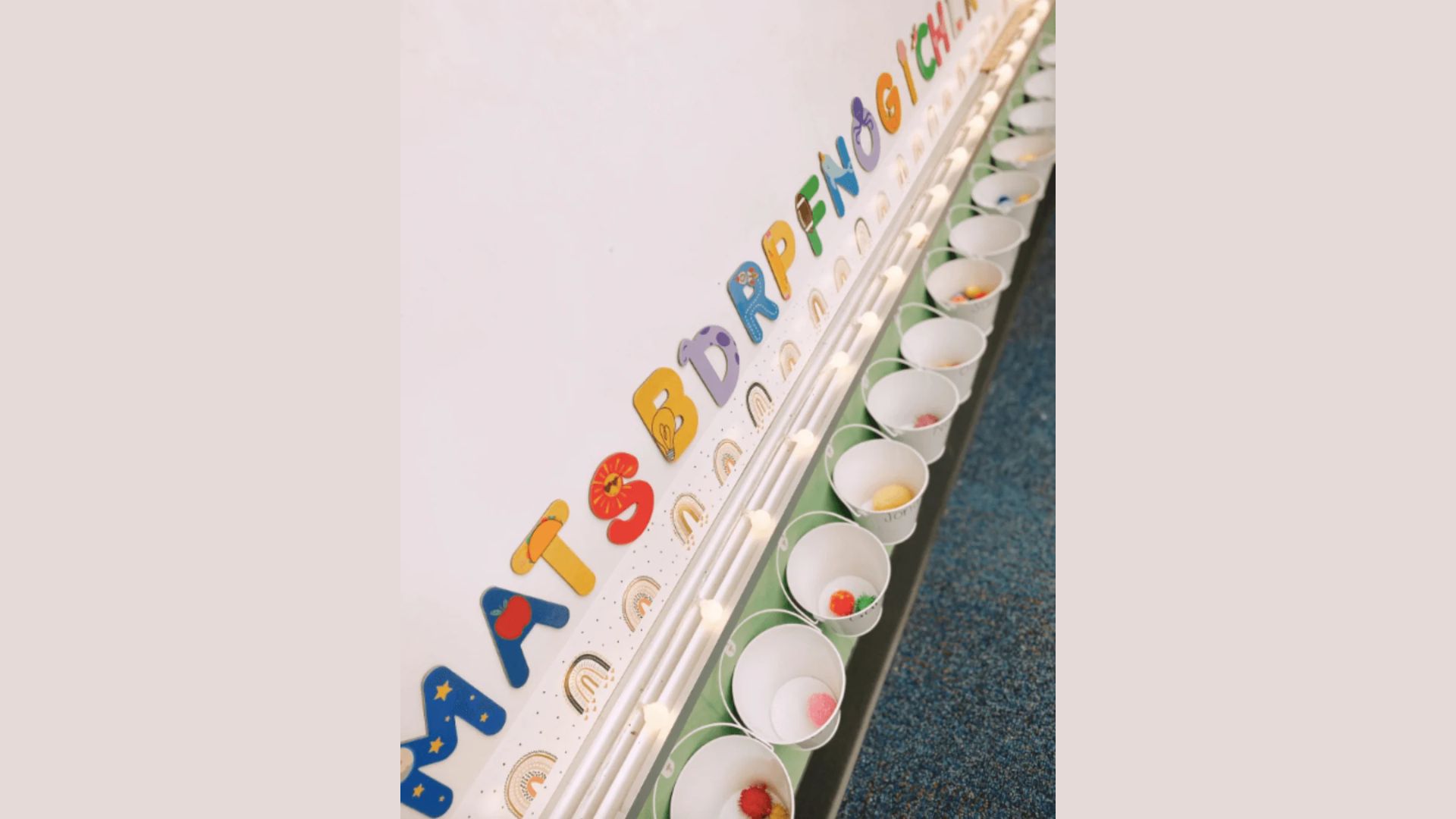 A row of small white buckets with student names lined up under alphabet letters on a classroom wall. Each bucket contains colorful pom-poms used as tokens to visually track kindness in this bucket filler activity for kids.