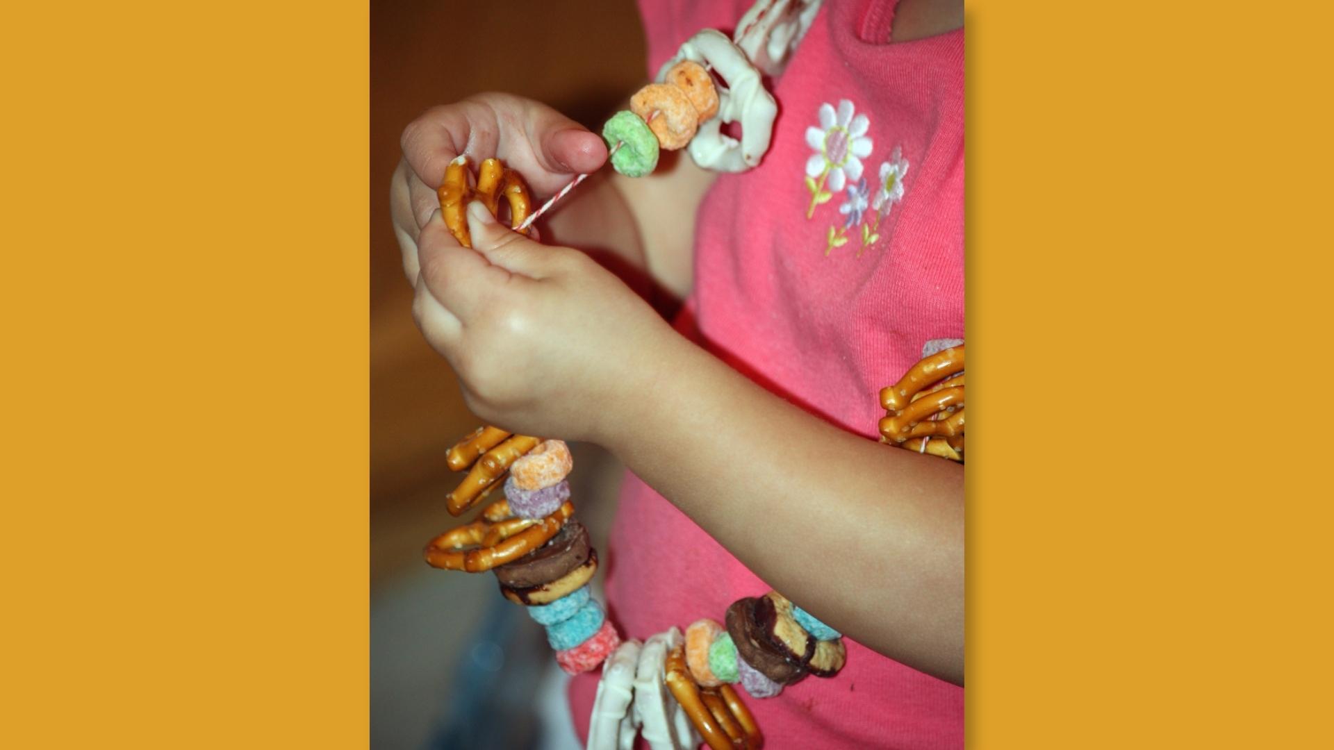 Child in pink shirt threads pretzels and cereal onto string making edible necklace.