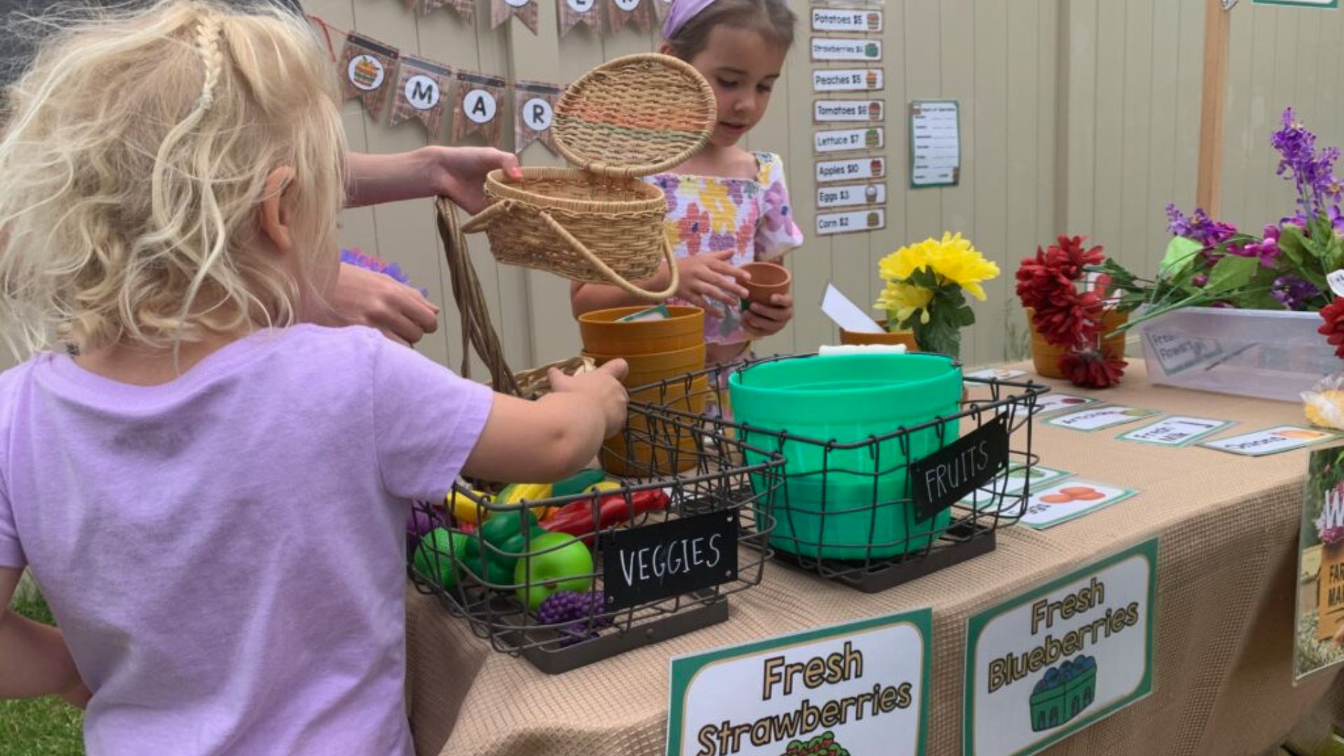 Two young girls playing pretend farmers market with toy fruits, vegetables, flowers, and price signs for fresh strawberries and blueberries, a dramatic play farm activity for preschoolers