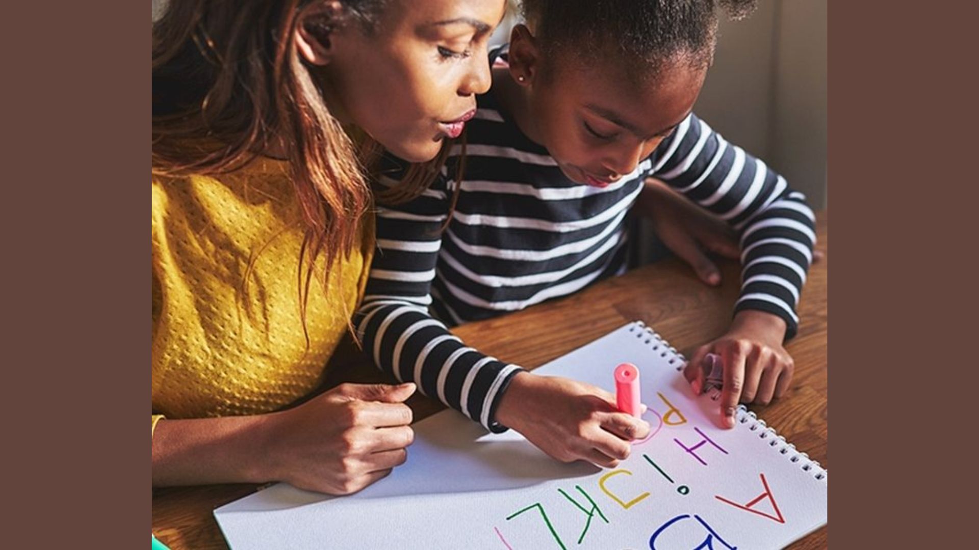An adult in a yellow sweater helps a child practice writing colorful alphabet letters in a notebook using a pink highlighter.