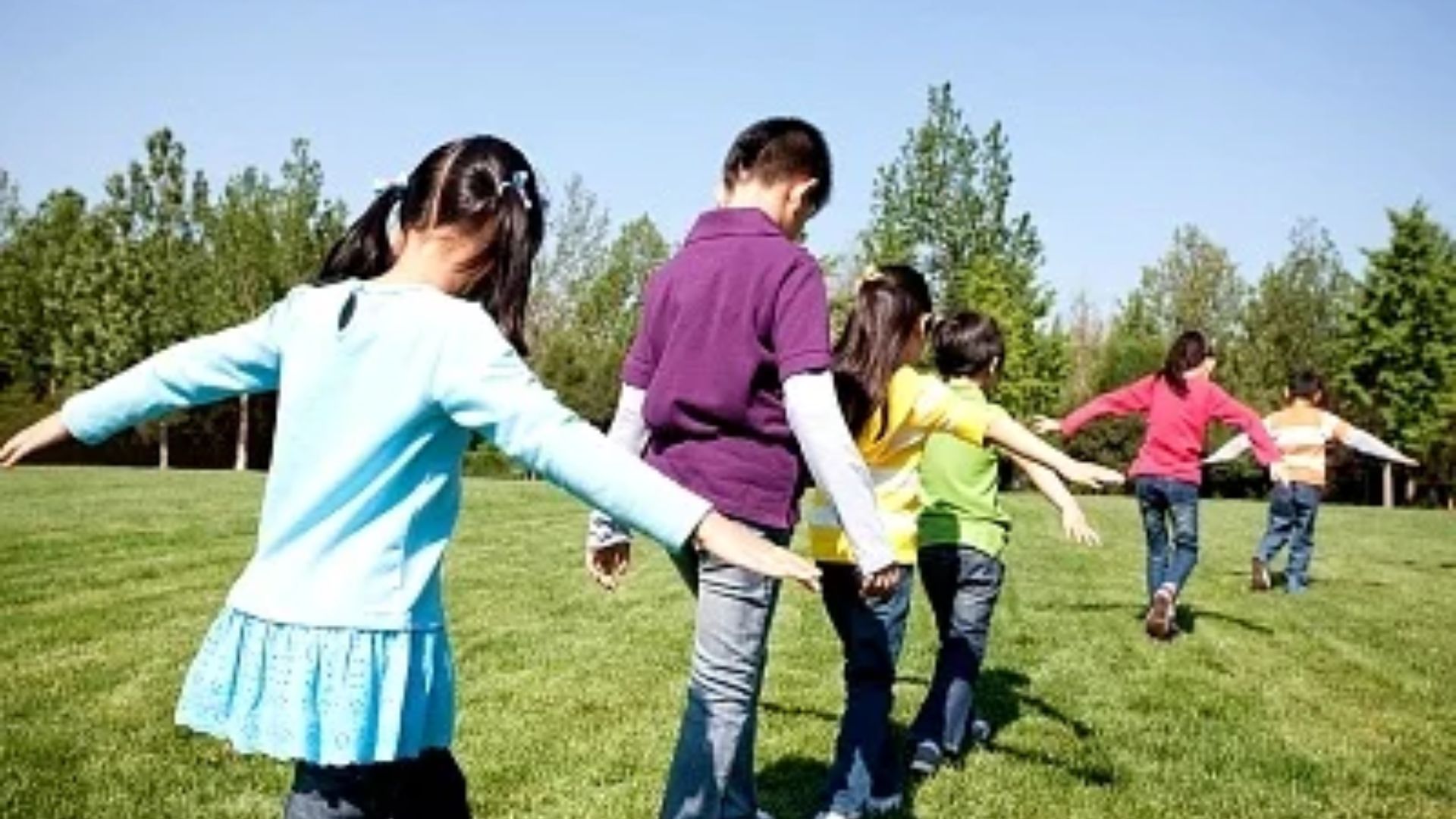 Line of diverse preschool children walking single-file on grass with arms out for balance, playing a cooperative follow-the-leader friendship activity for preschoolers