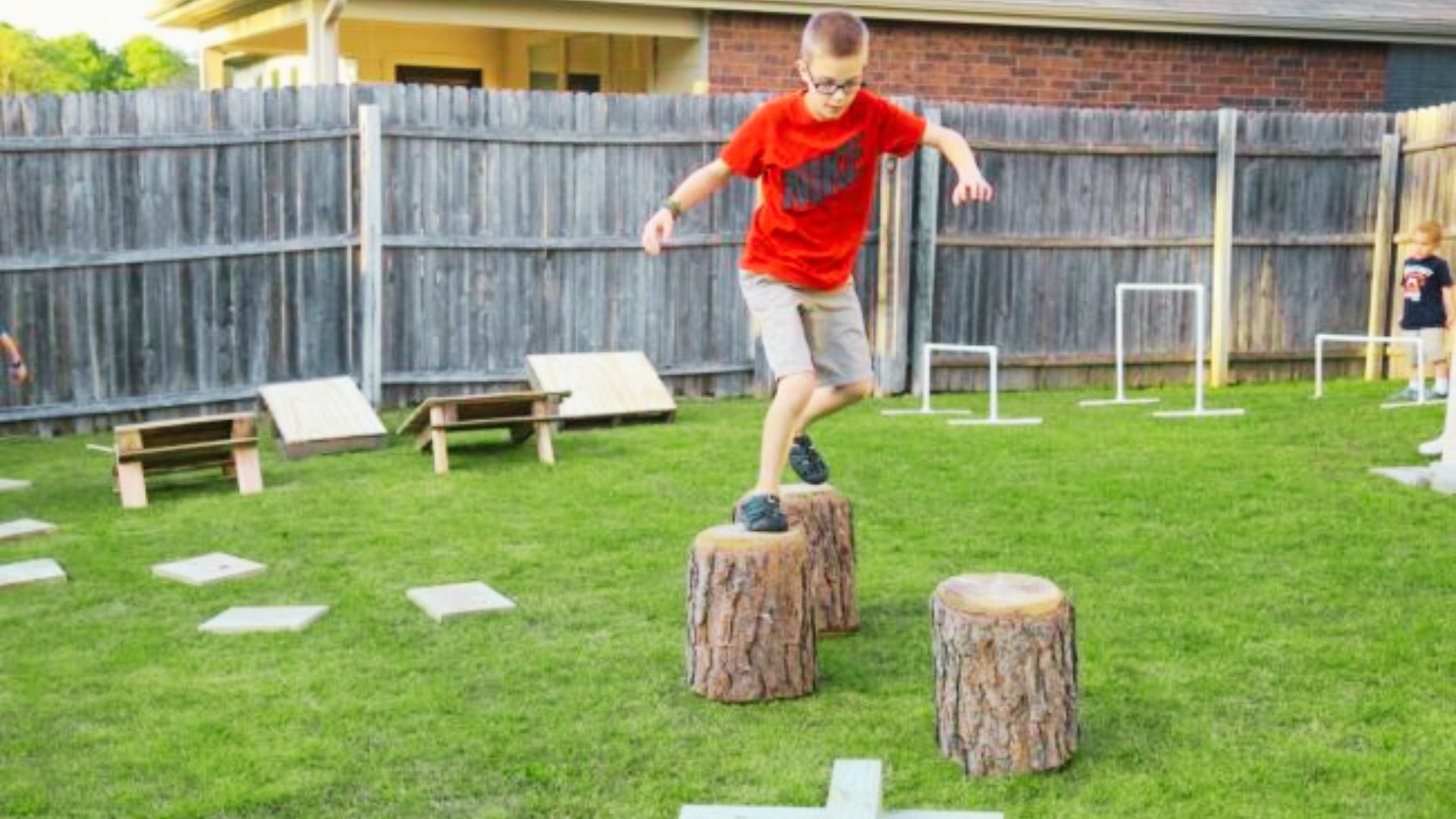 A boy joyfully jumps over logs in a sunny backyard, showcasing his playful spirit and energy.