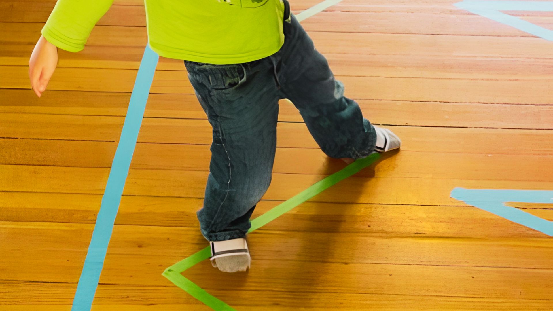 A young boy in a green shirt stands on a wooden floor with tape on it.