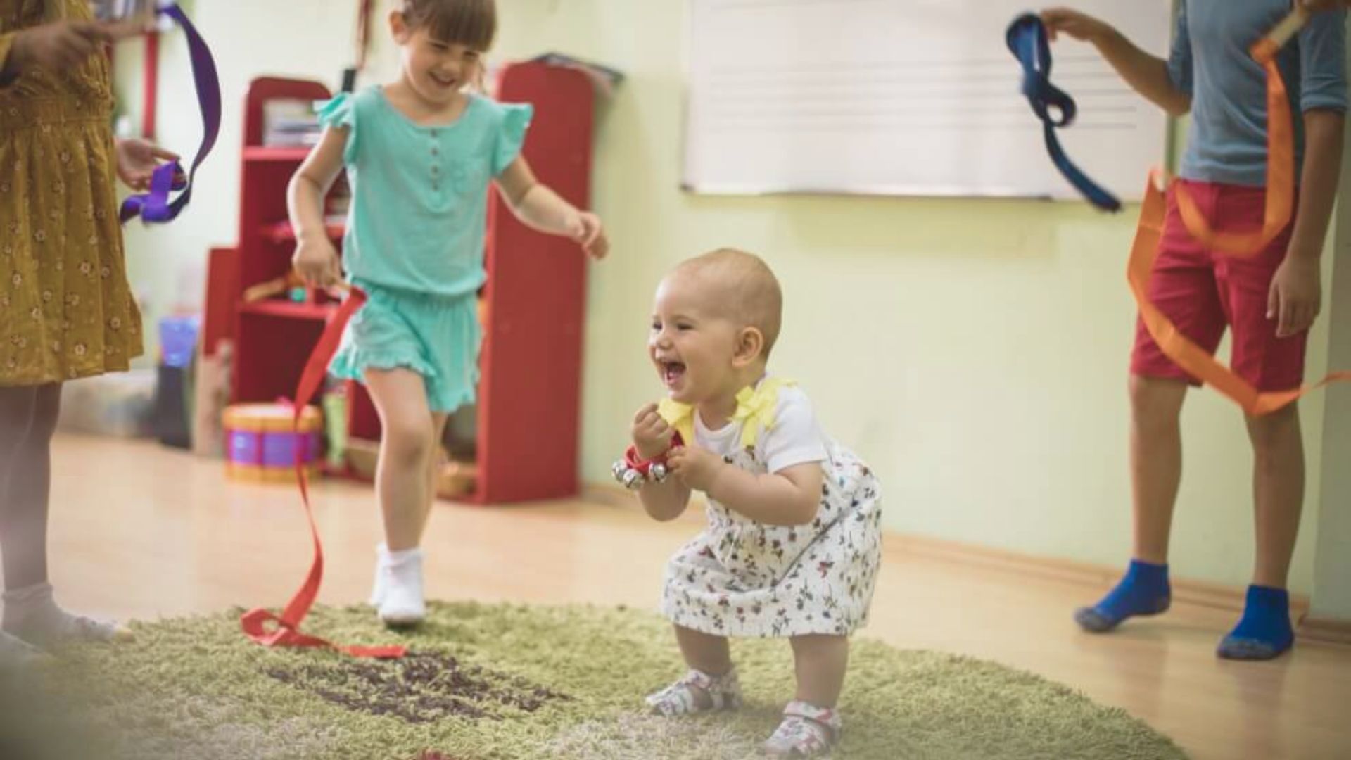 A laughing baby in a floral dress stands on a rug holding jingle bells while older children wave ribbons.