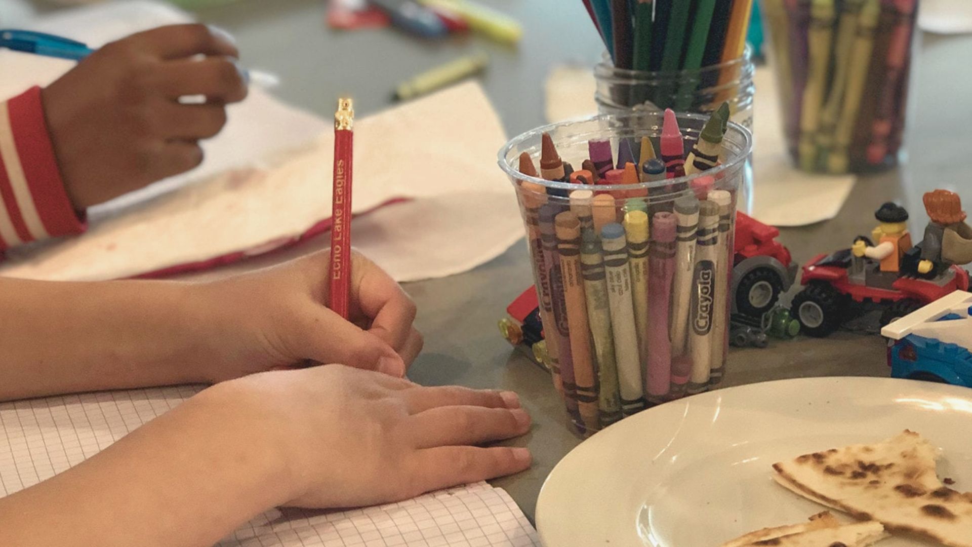 A child’s hand holds a red pencil while drawing on graph paper, surrounded by crayons and toys.