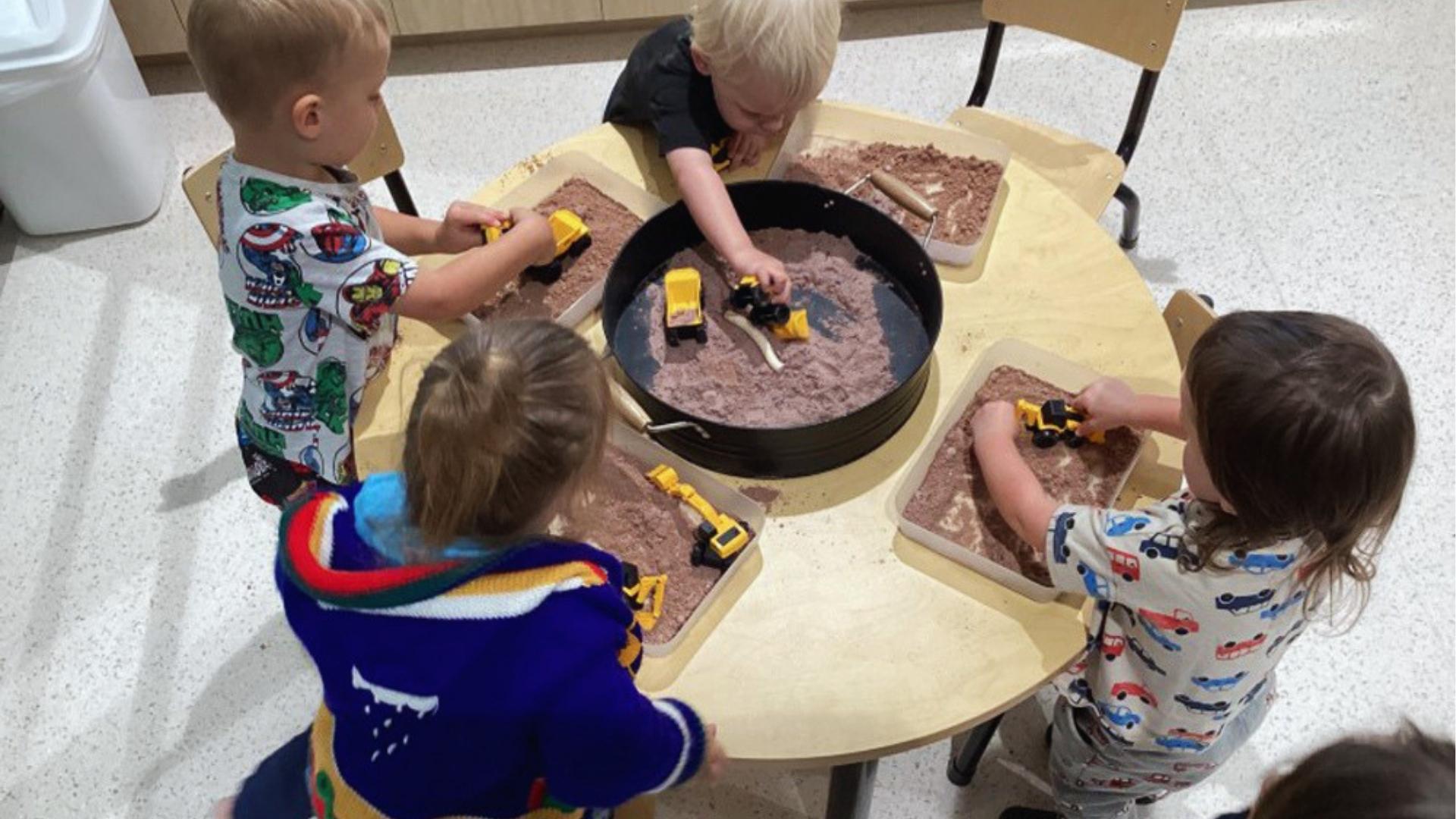 Preschoolers play with toy trucks in sand trays together