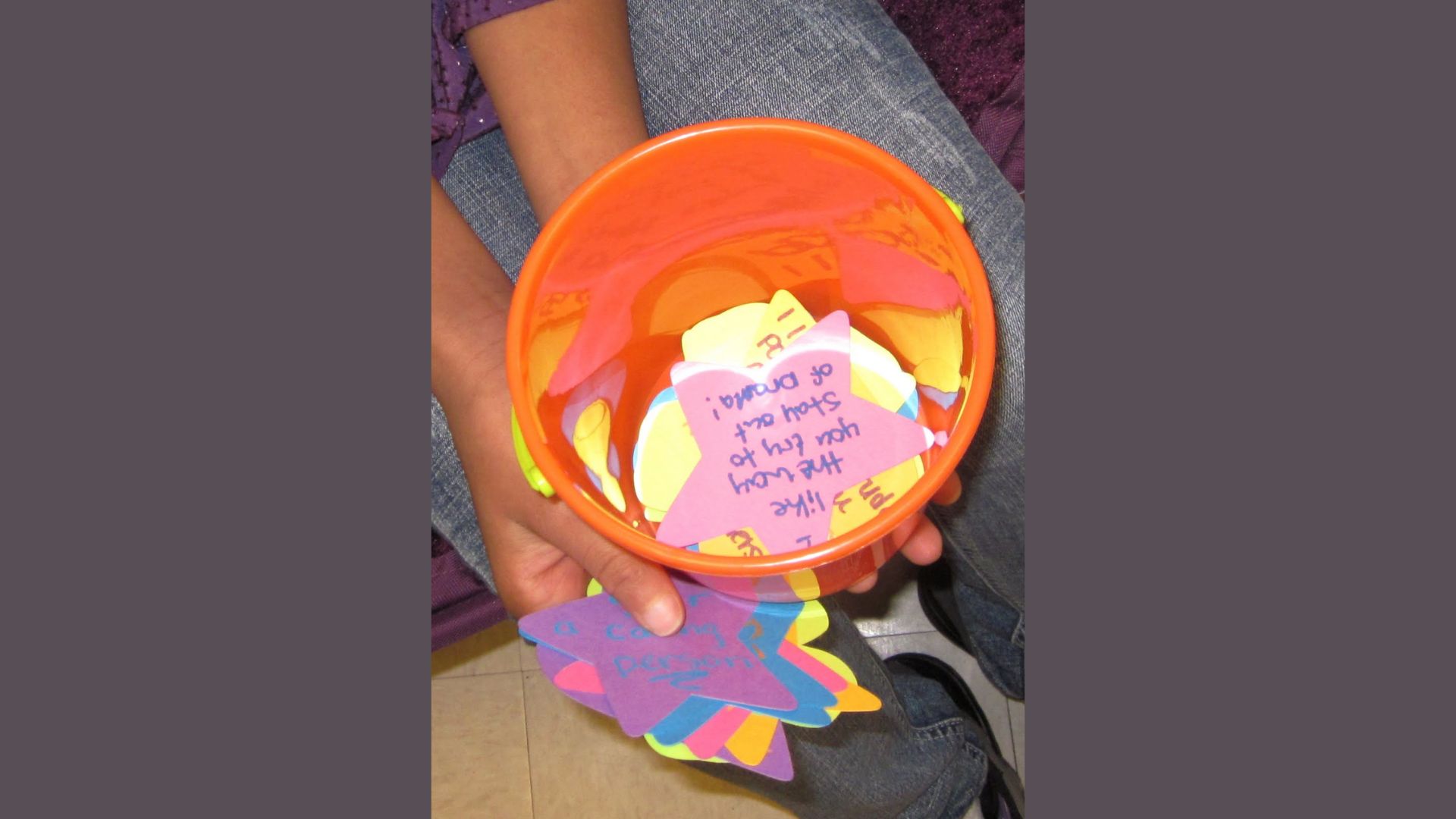 Child’s hands holding an orange plastic bucket filled with colorful paper stars and hearts with handwritten kindness notes.