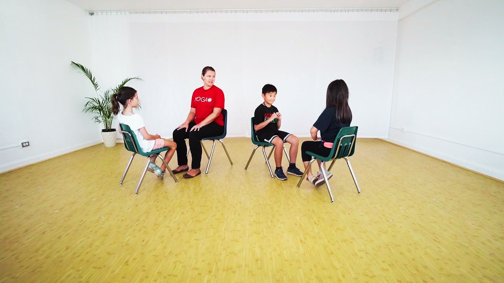 children seated in chairs, engaging in chair yoga stretches in a spacious, empty room.
