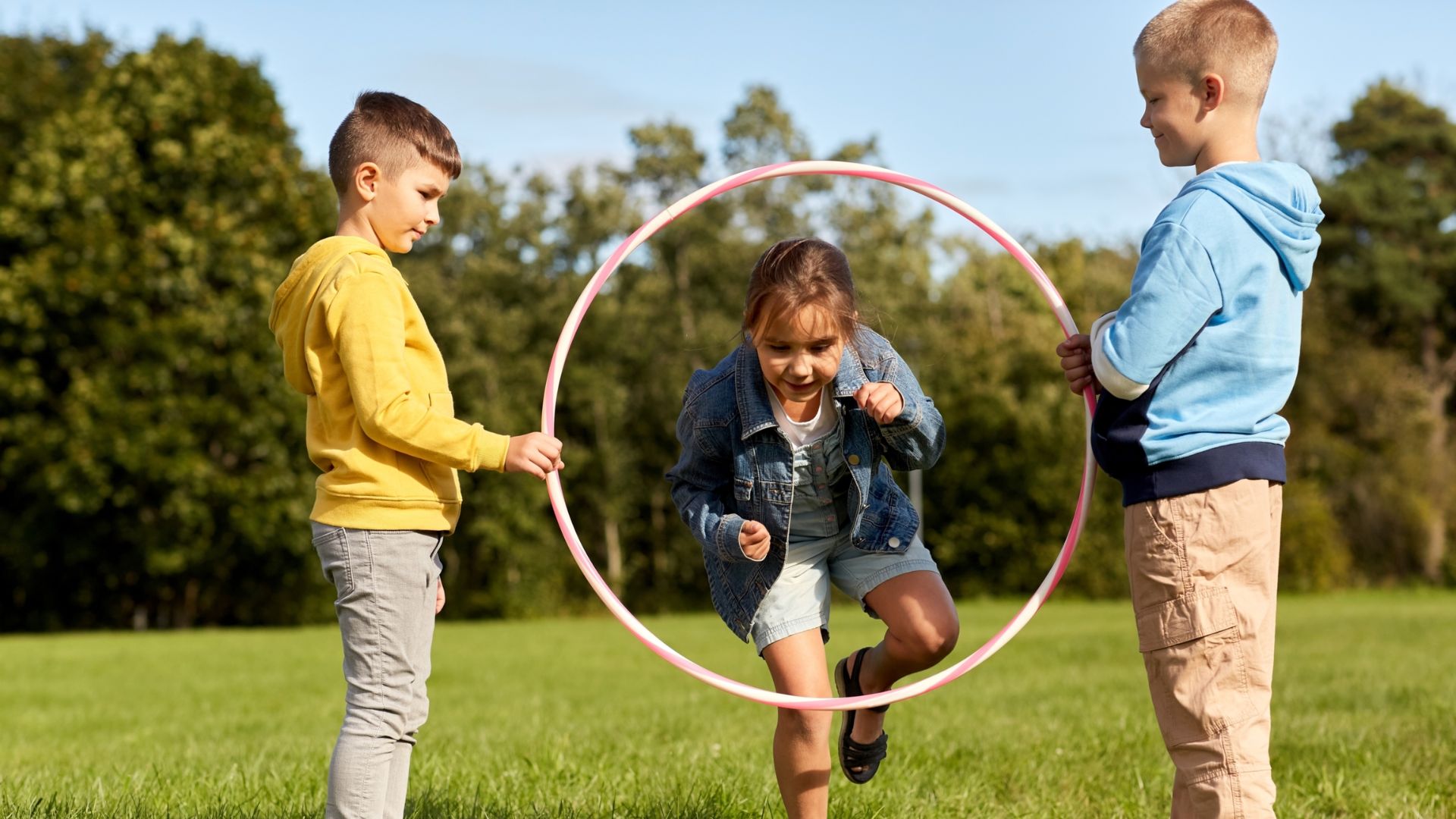 Three children joyfully playing with a hula hoop in a grassy field during a personal space activity.