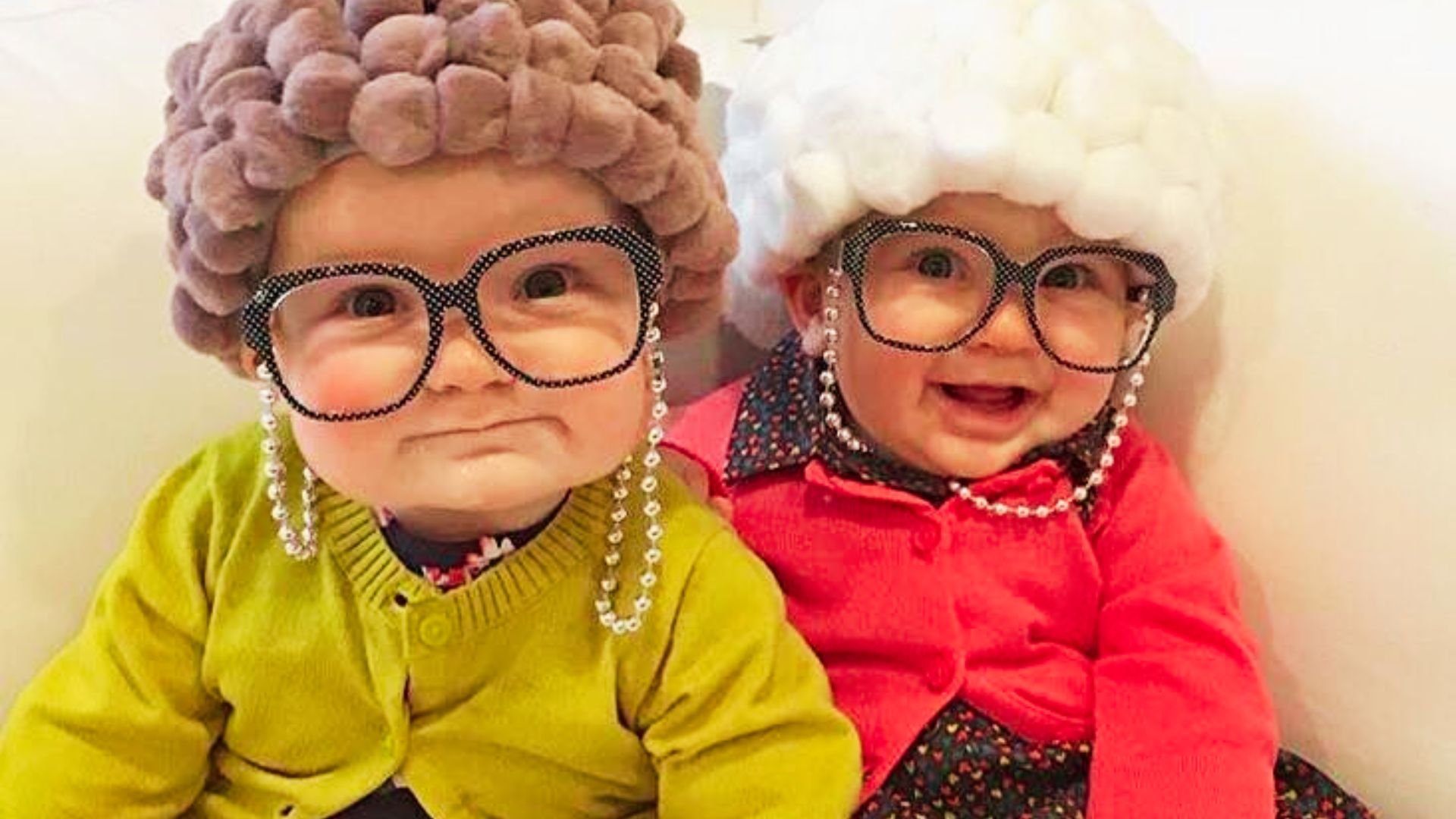 Two babies dressed as elderly women, complete with glasses and shawls, sitting together and smiling.