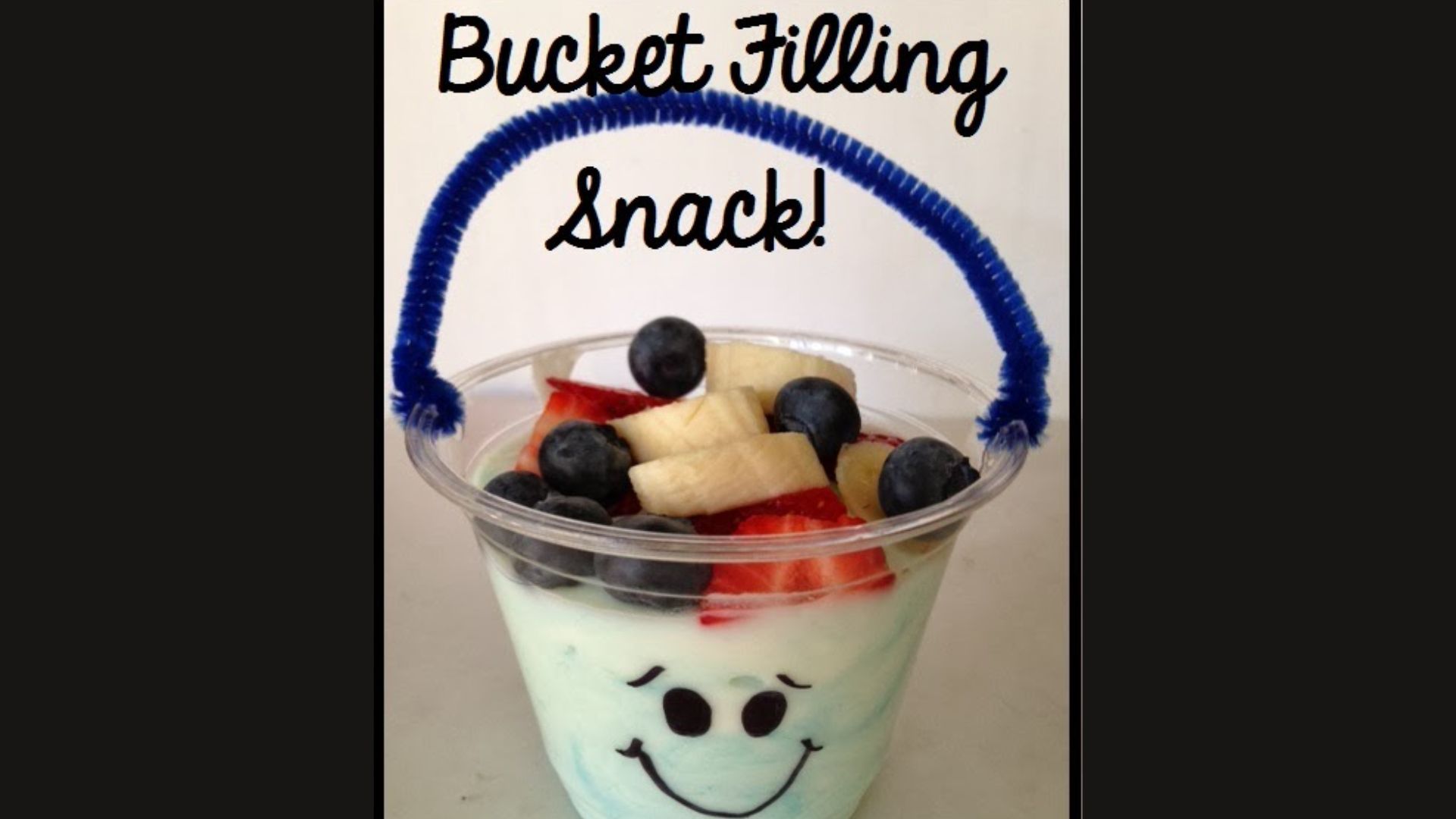 “Bucket Filling Snack” in a clear plastic cup decorated with a smiley face and blue pipe cleaner handle, filled with yogurt, strawberries, blueberries, and banana slices.