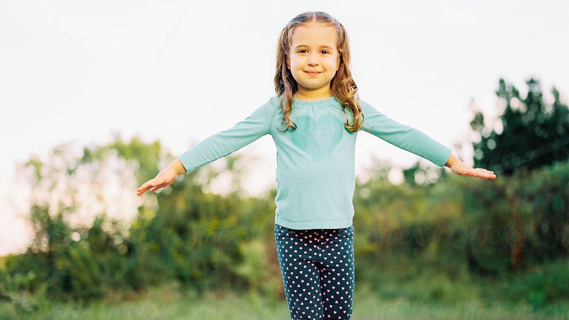 A little girl stands on one leg in a grassy area, smiling and balancing with her arms outstretched.