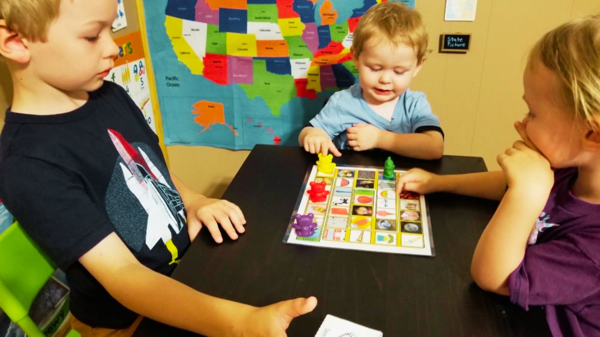 Three young children engaged in a board game, smiling and focused, seated around a table in a bright room.