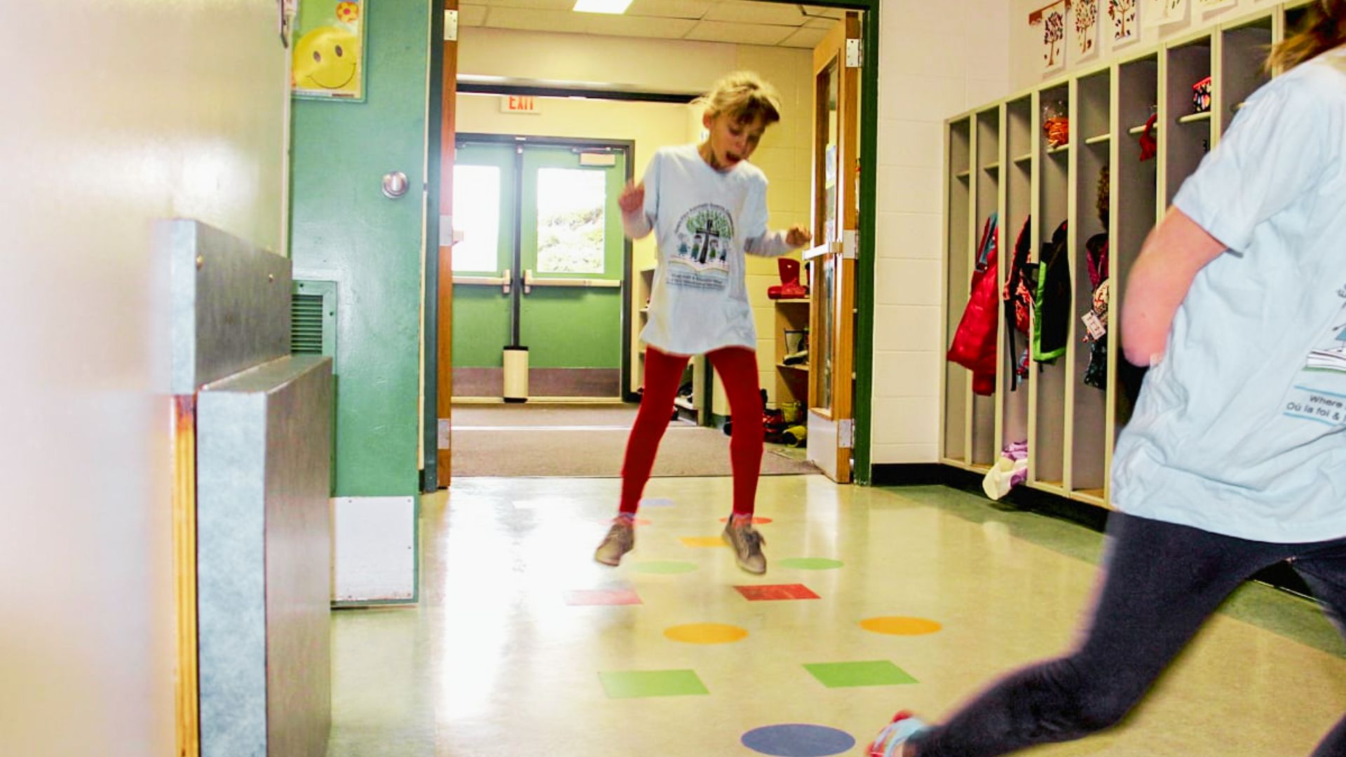 A girl joyfully playing hopscotch in a brightly lit hallway, with chalk markings on the floor.
