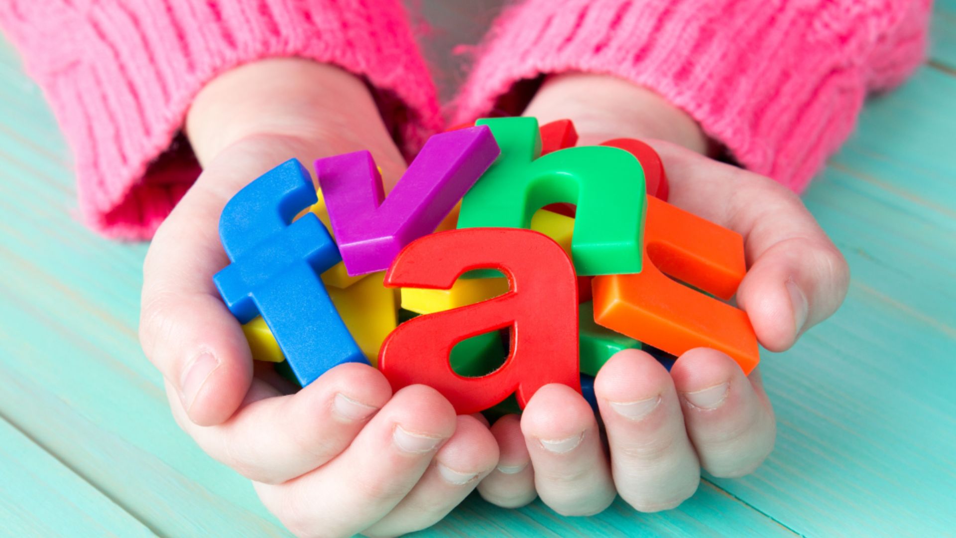 A child’s hands in a pink sweater hold a pile of bright, multicolored plastic alphabet letters including f, y, a, and d.