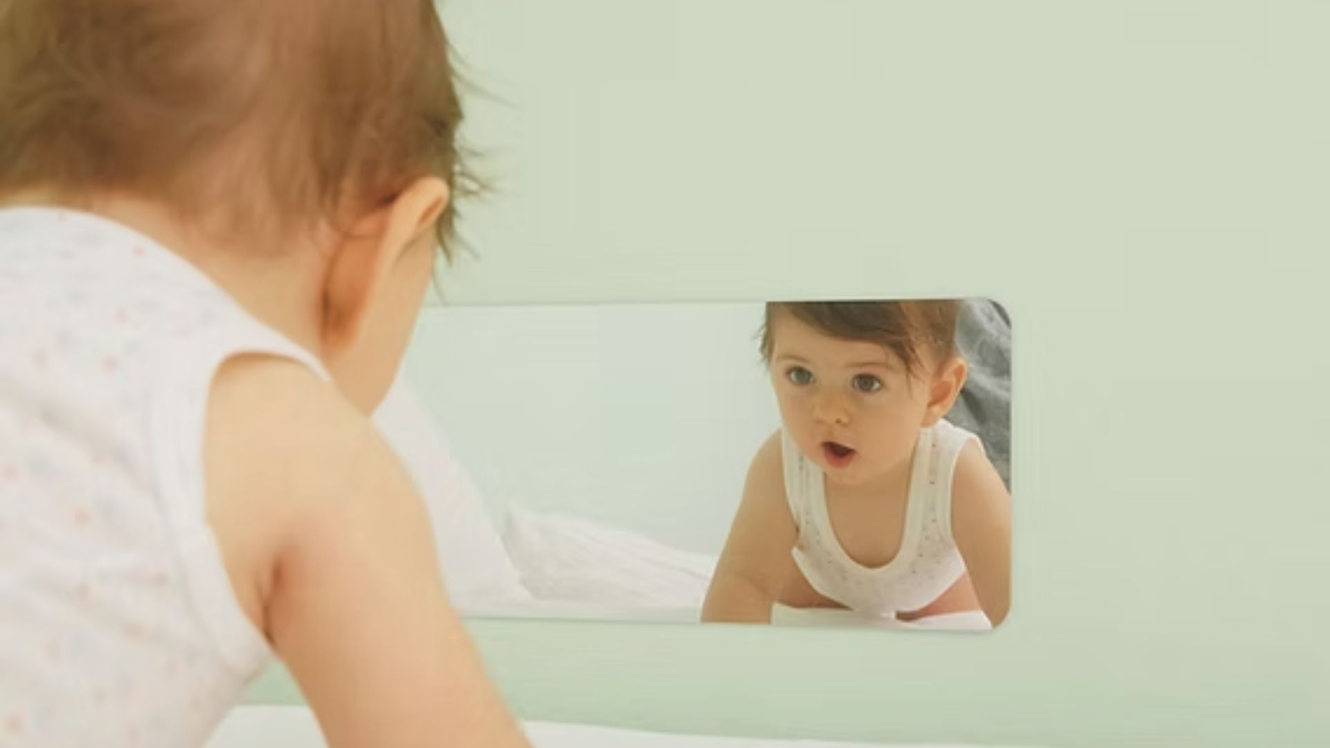 A baby in a white onesie looks at their reflection in a low mirror