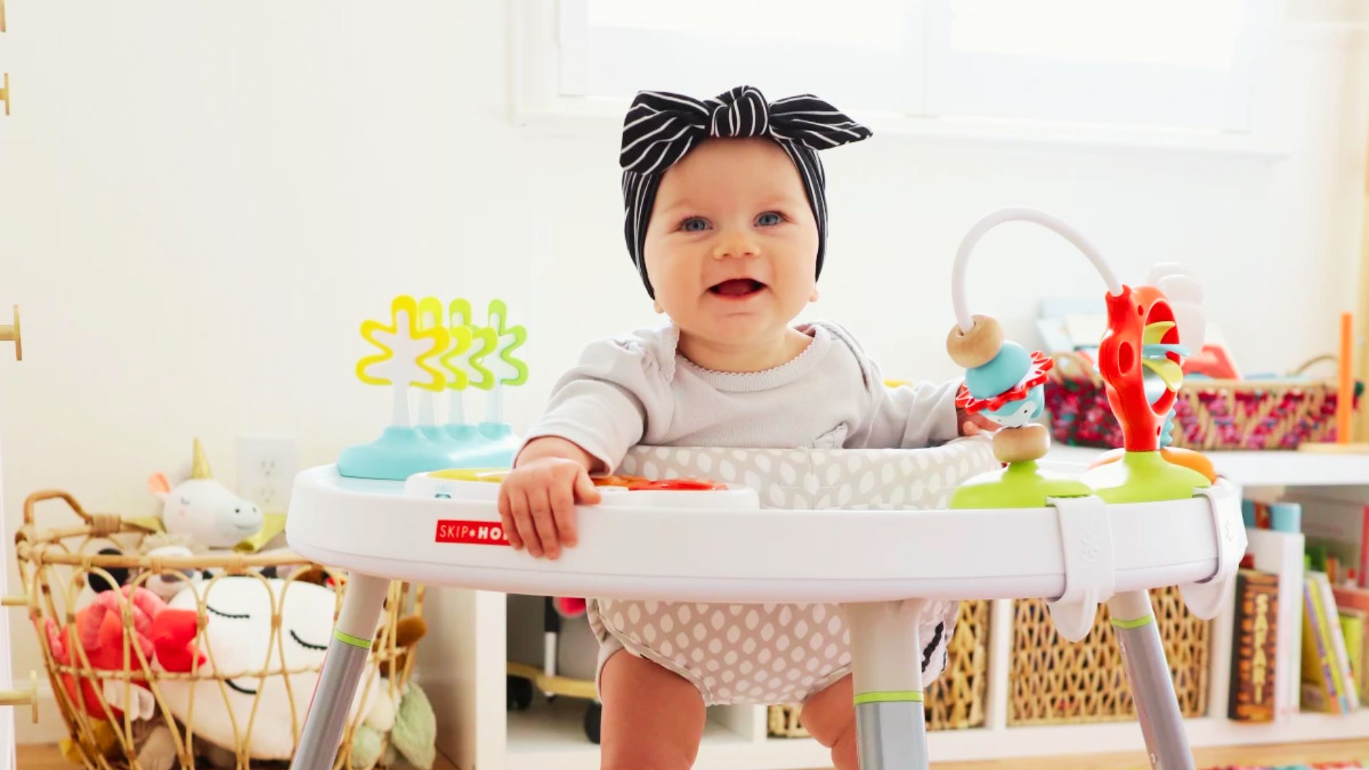 A baby in a walker, wearing a bow on her head, smiles while finding her surroundings.