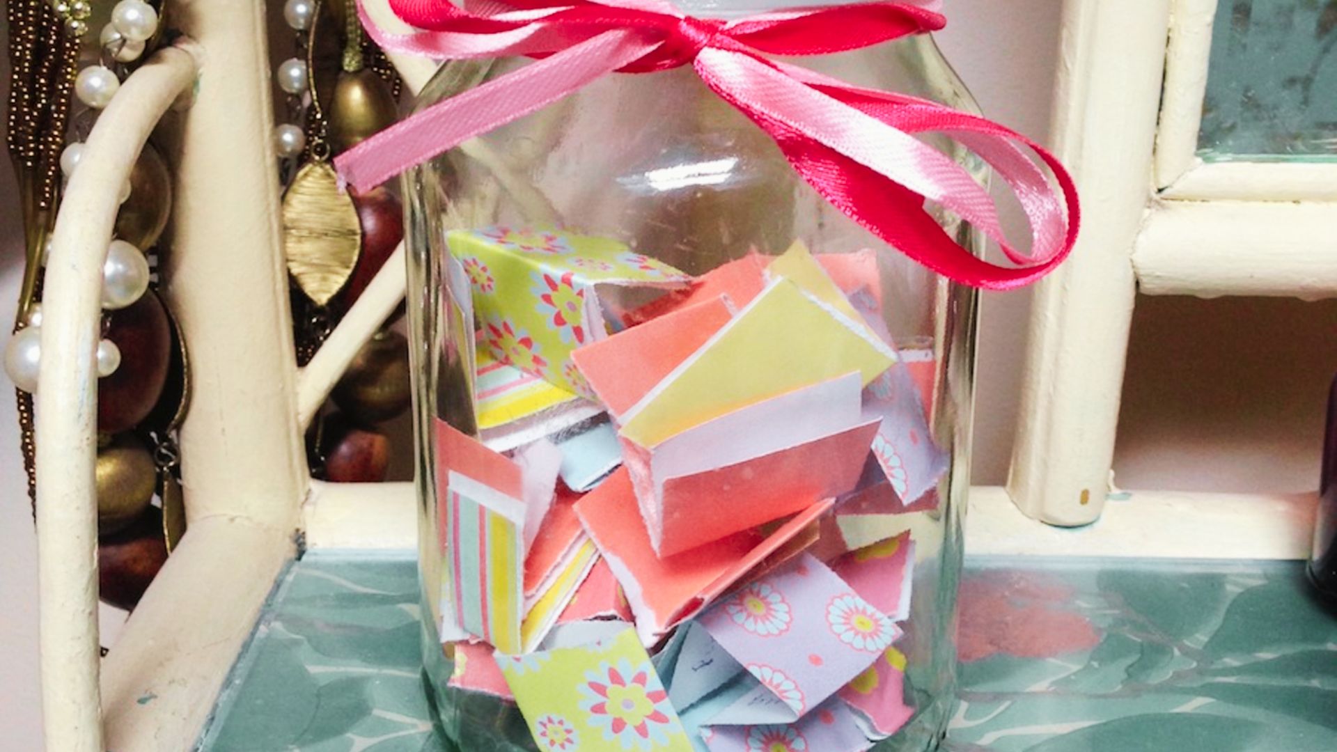 A jar filled with colorful paper scraps, tied with a pink ribbon, sitting on a wooden surface.