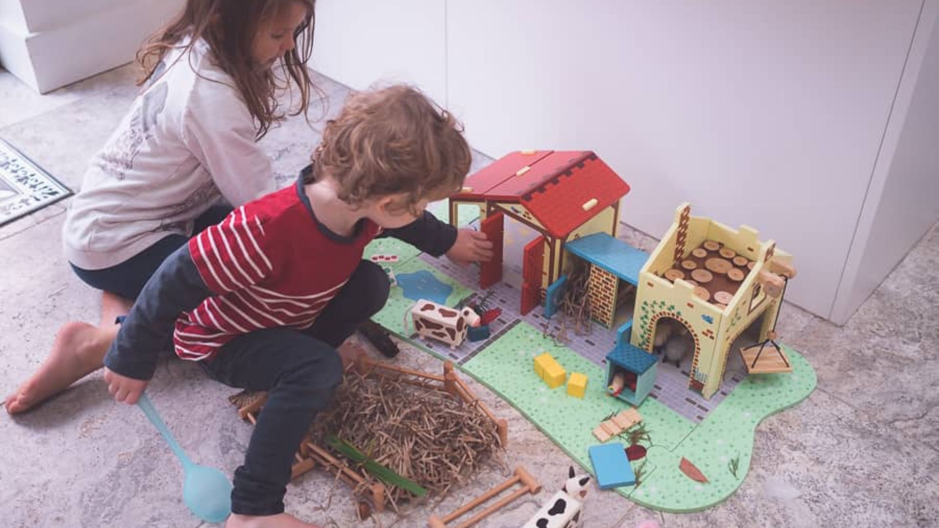 Two children playing with a wooden toy farm set including barn, animals, hay, and fences, a dramatic play farm activity for home