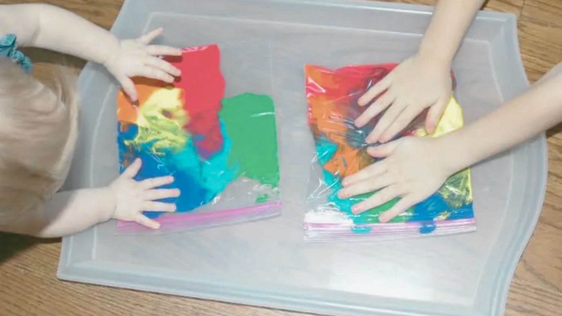 Two sets of small hands press and smear colorful paint inside sealed ziplock bags on a tray.