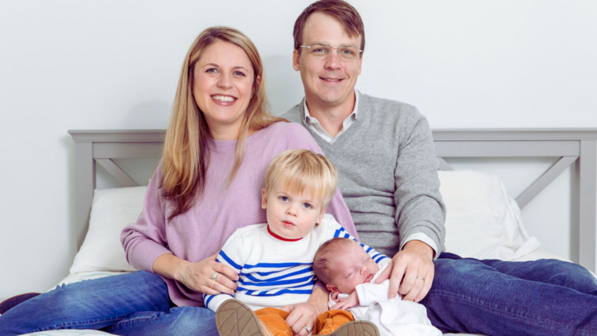 A man and woman sit on a bed, smiling at their baby who is lying between them, creating a warm family moment.
