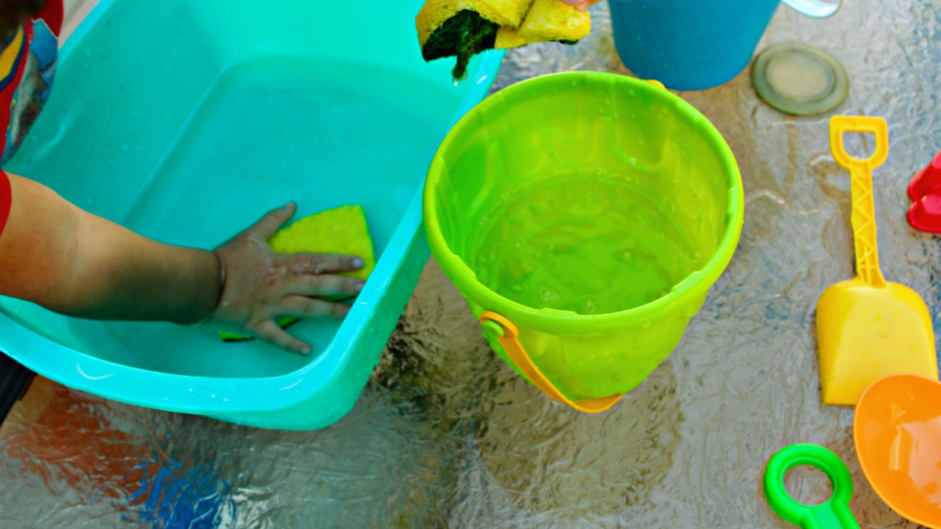 A toddlerโs hands press a yellow sponge in a blue tub while water drips into a green bucket nearby.