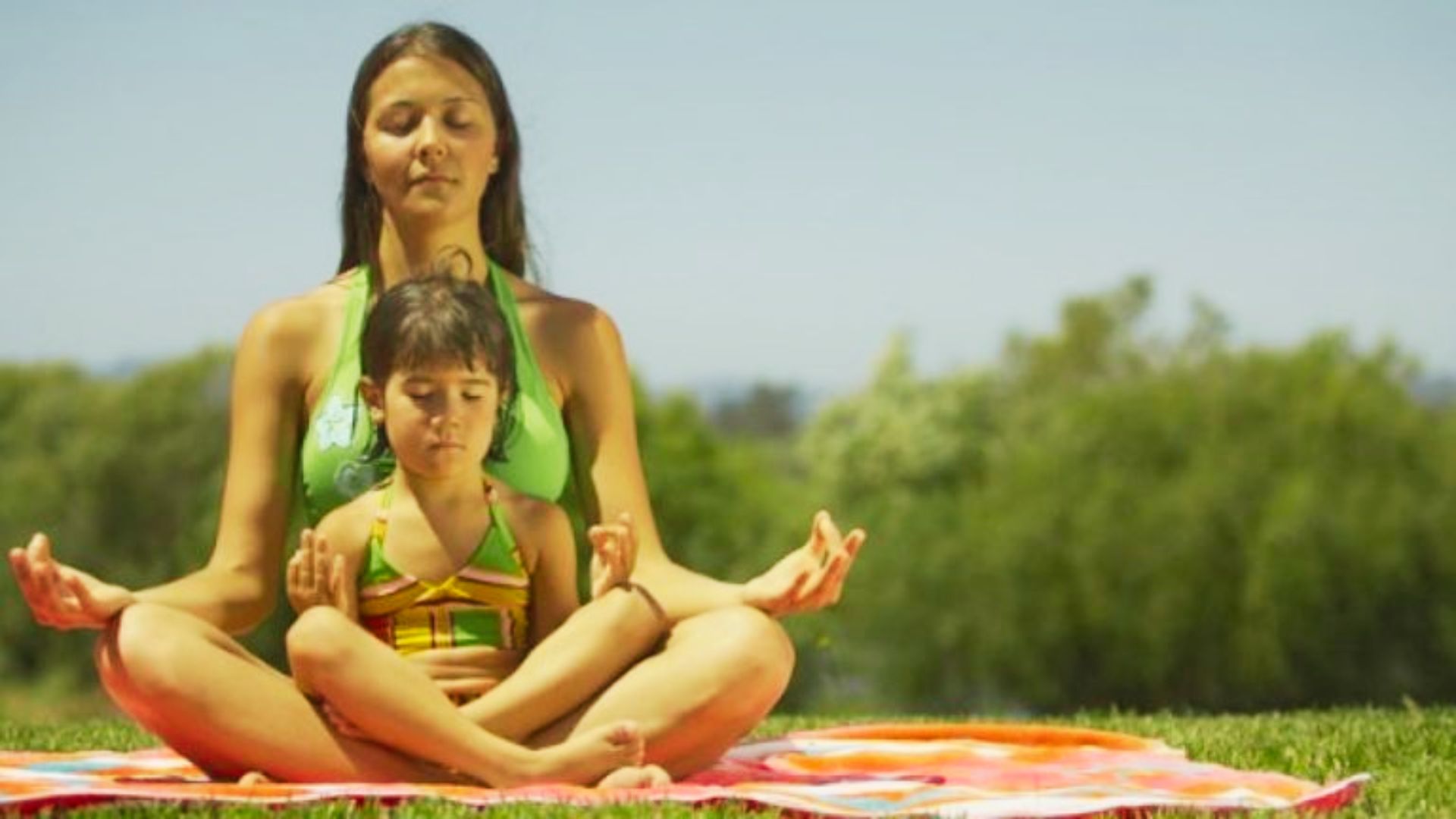 A woman and a child sit cross-legged on the grass, peacefully meditating together in a serene outdoor setting.