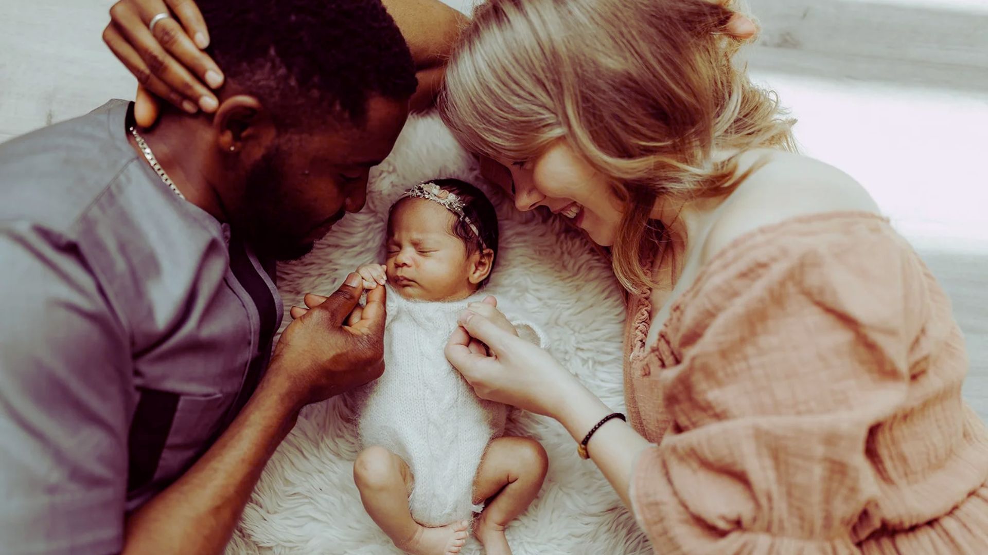 A couple holds their baby girl while a man gazes at her with a warm smile, capturing a moment of family affection.