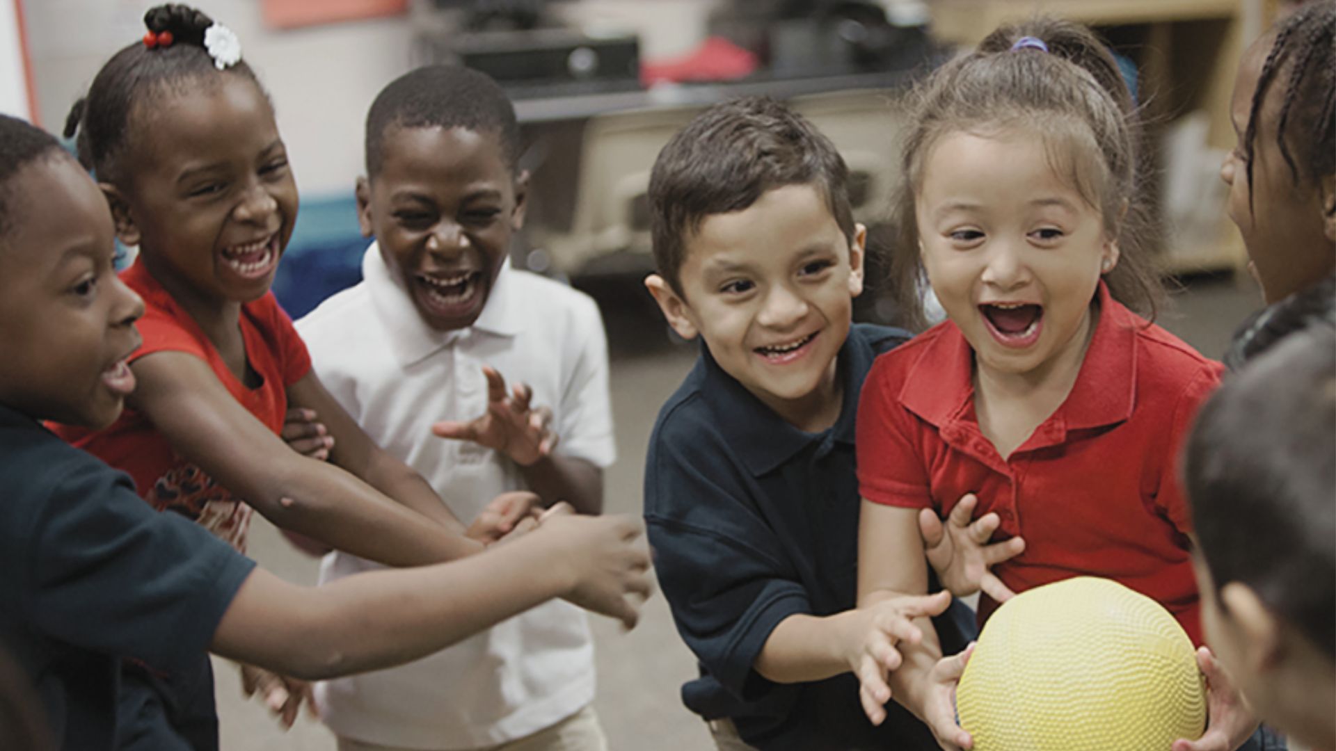 Excited group of diverse preschoolers laughing and reaching for a yellow ball in a circle, practicing cooperative play and social engagement in a friendship activity