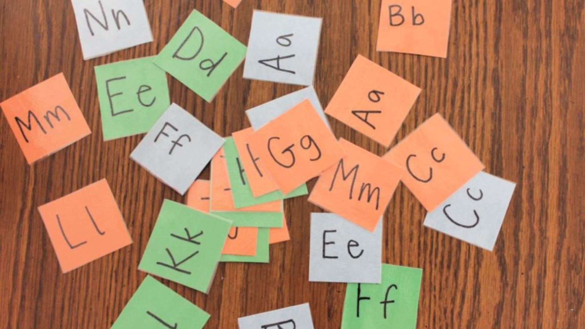 Scattered orange, green, and white paper squares with handwritten uppercase and lowercase letter pairs like Aa, Bb, Cc on wood surface.
