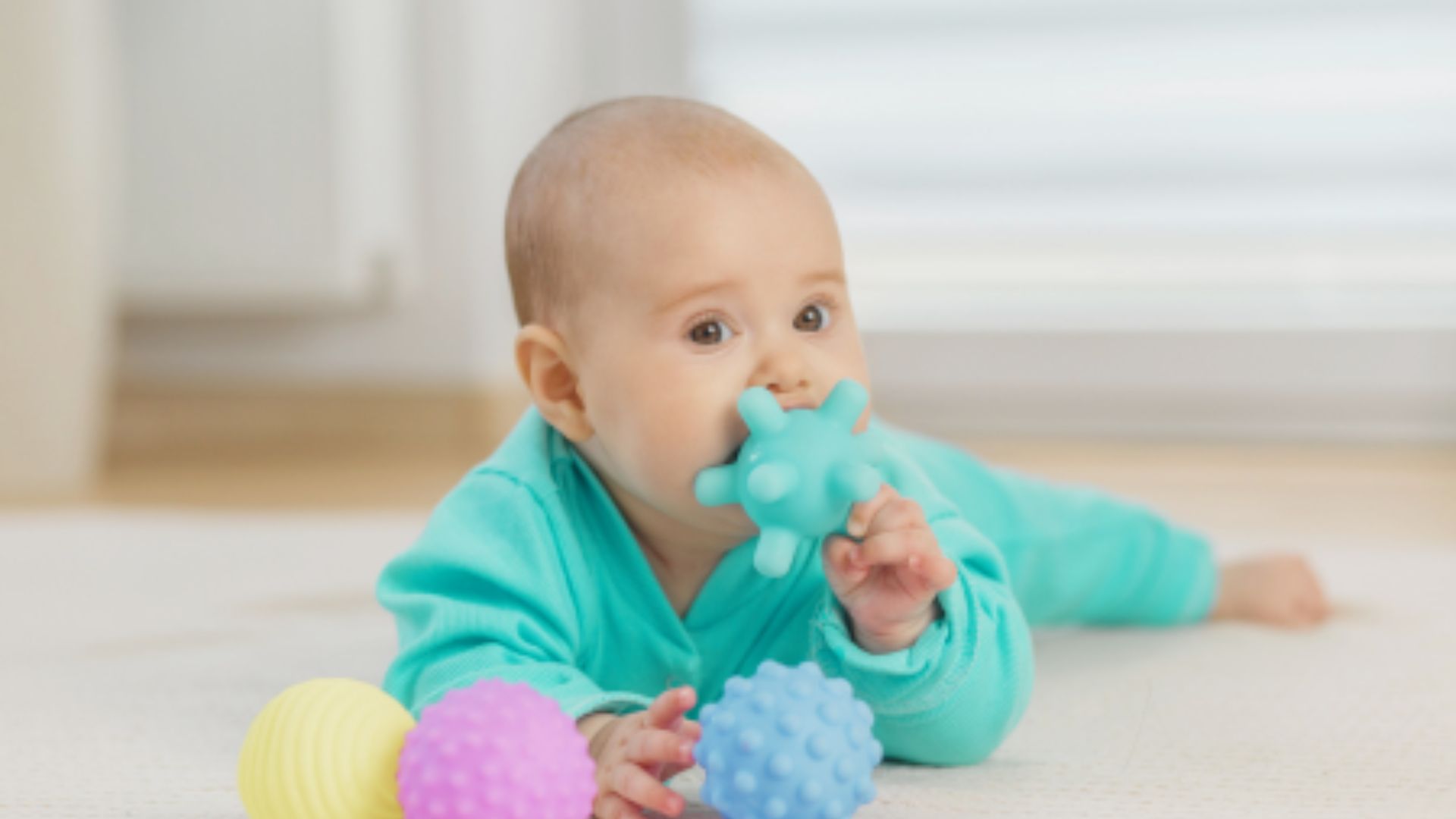 An infant in a turquoise onesie lies on their tummy, mouthing a textured blue sensory toy during playtime.