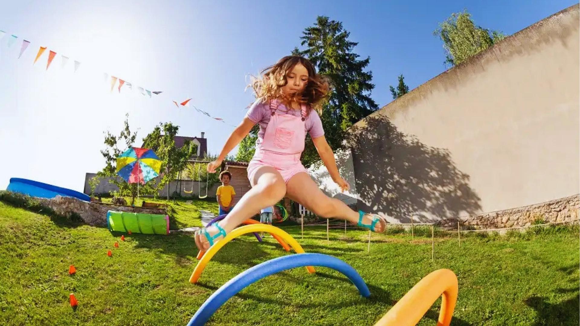 Girl in pink overalls jumps over pool noodle hurdles on grass, flags, sunny, Summer Activities for Kids.