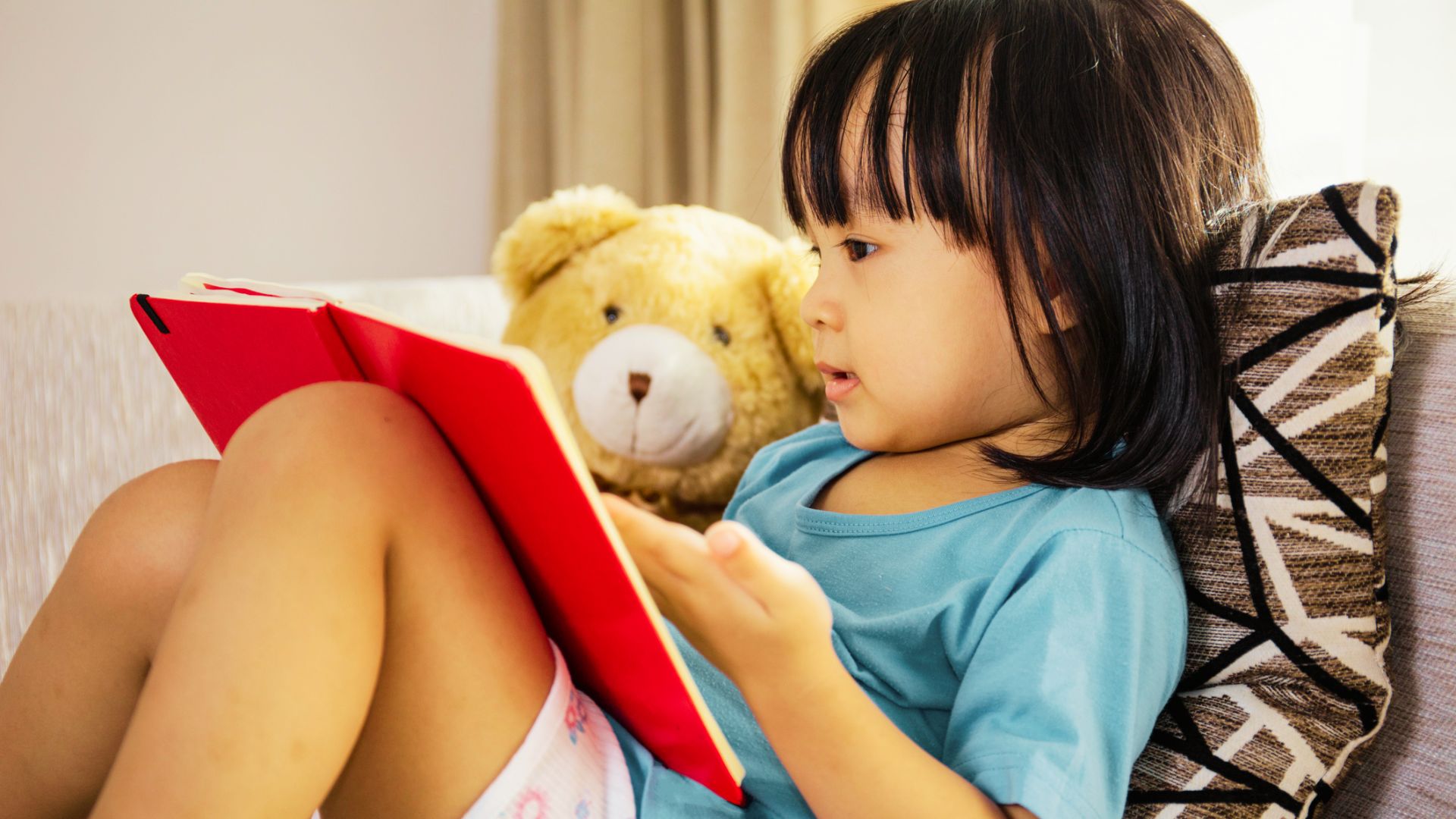 A little girl sits on a couch, holding a teddy bear, with a joyful expression on her face.