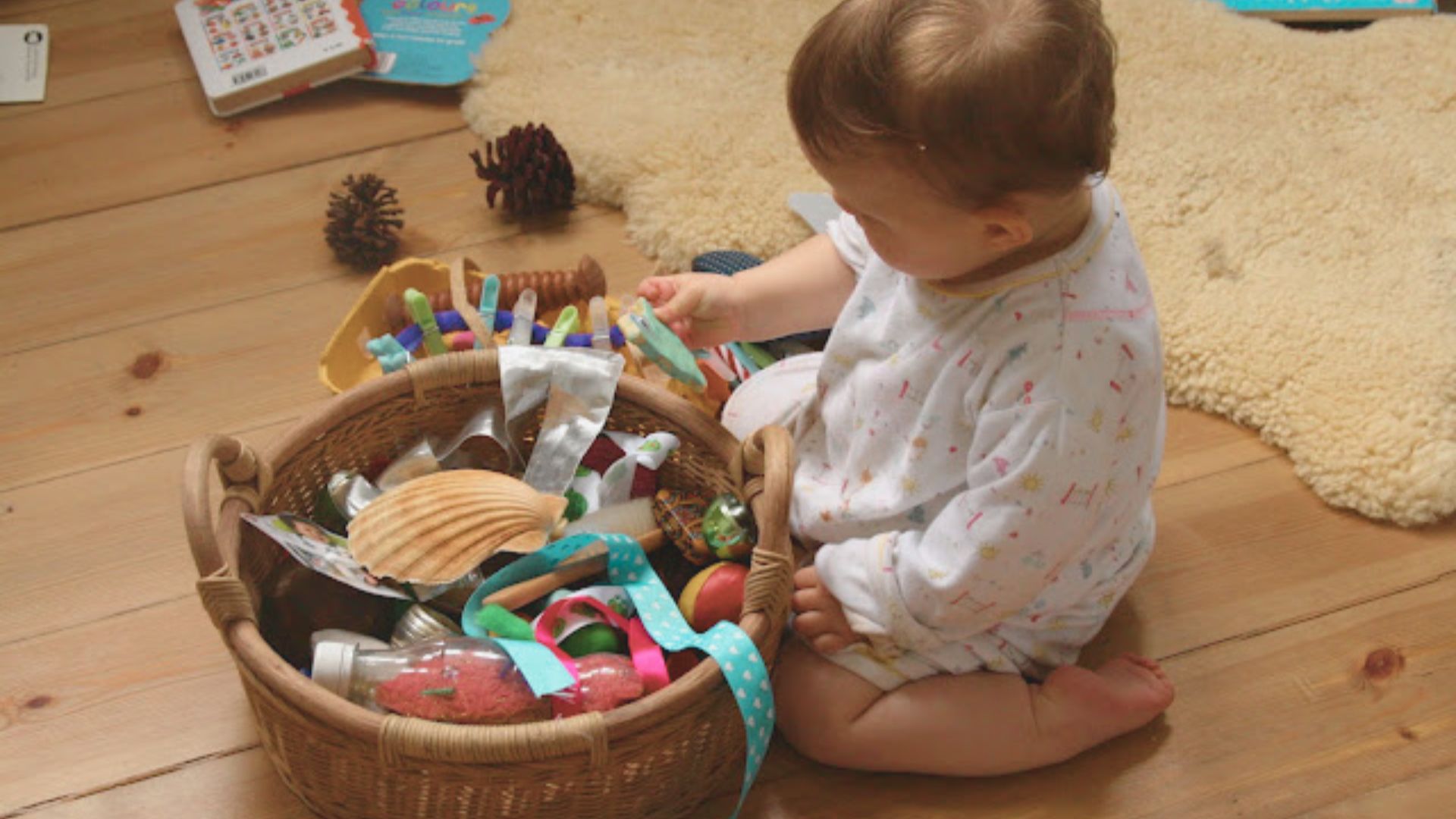 An infant in a white onesie sits on a wooden floor and finds objects from a wicker treasure basket.