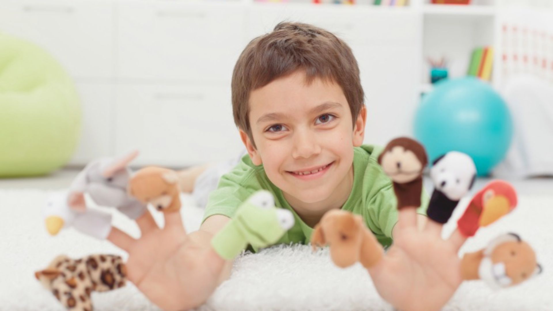 A smiling boy lies on a carpet showing animal finger puppets on both hands.