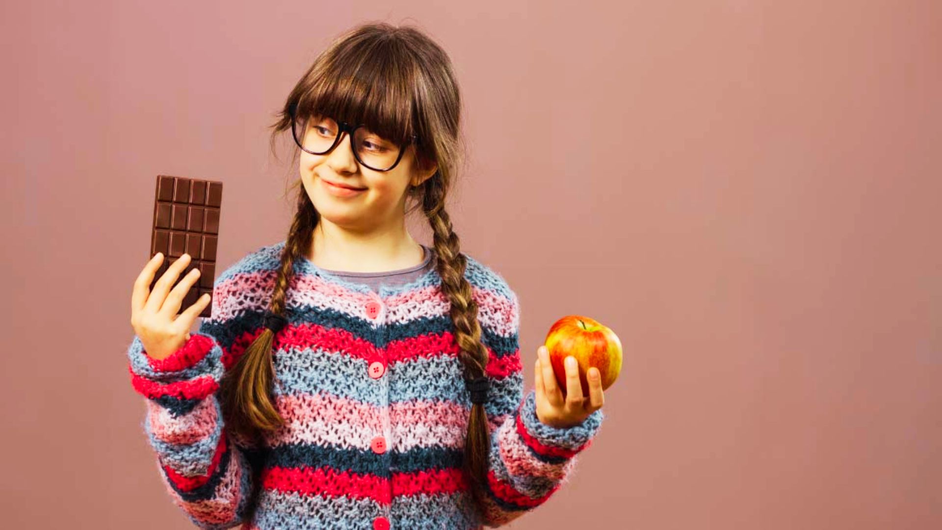 A girl with glasses holds an apple in one hand and a chocolate bar in the other, smiling at the camera.