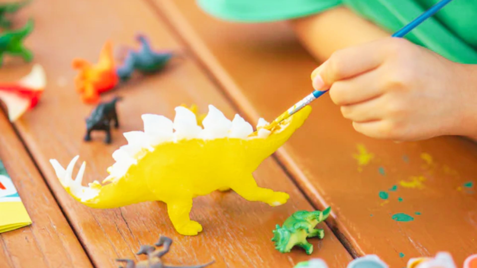 A child paints a toy dinosaur with a brush, focusing intently on adding color to its scales.