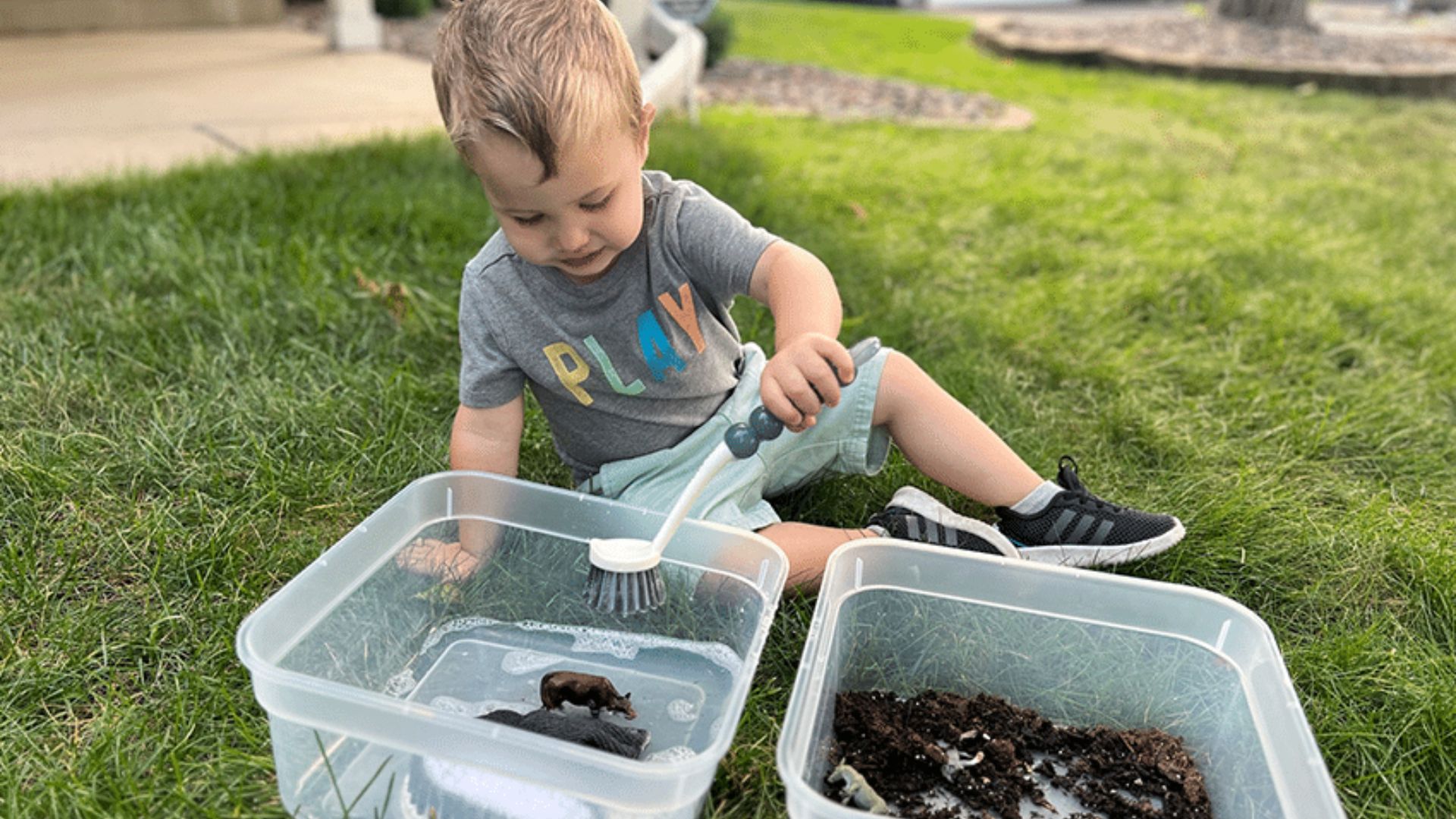 Toddler sitting on grass using a brush to wash muddy toy farm animals in a bin of soapy water next to a bin of dirt, a sensory play farm activity for home and classroom