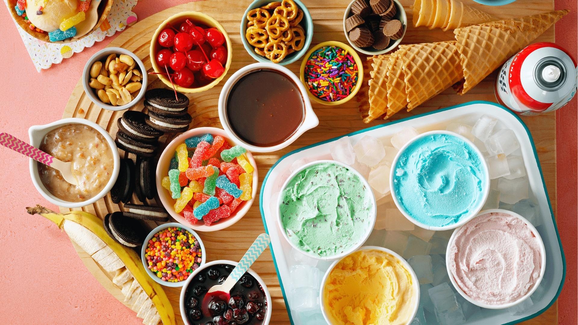 Ice cream sundae bar with toppings, sprinkles, cherries, cones, sauces, four ice cream flavors.