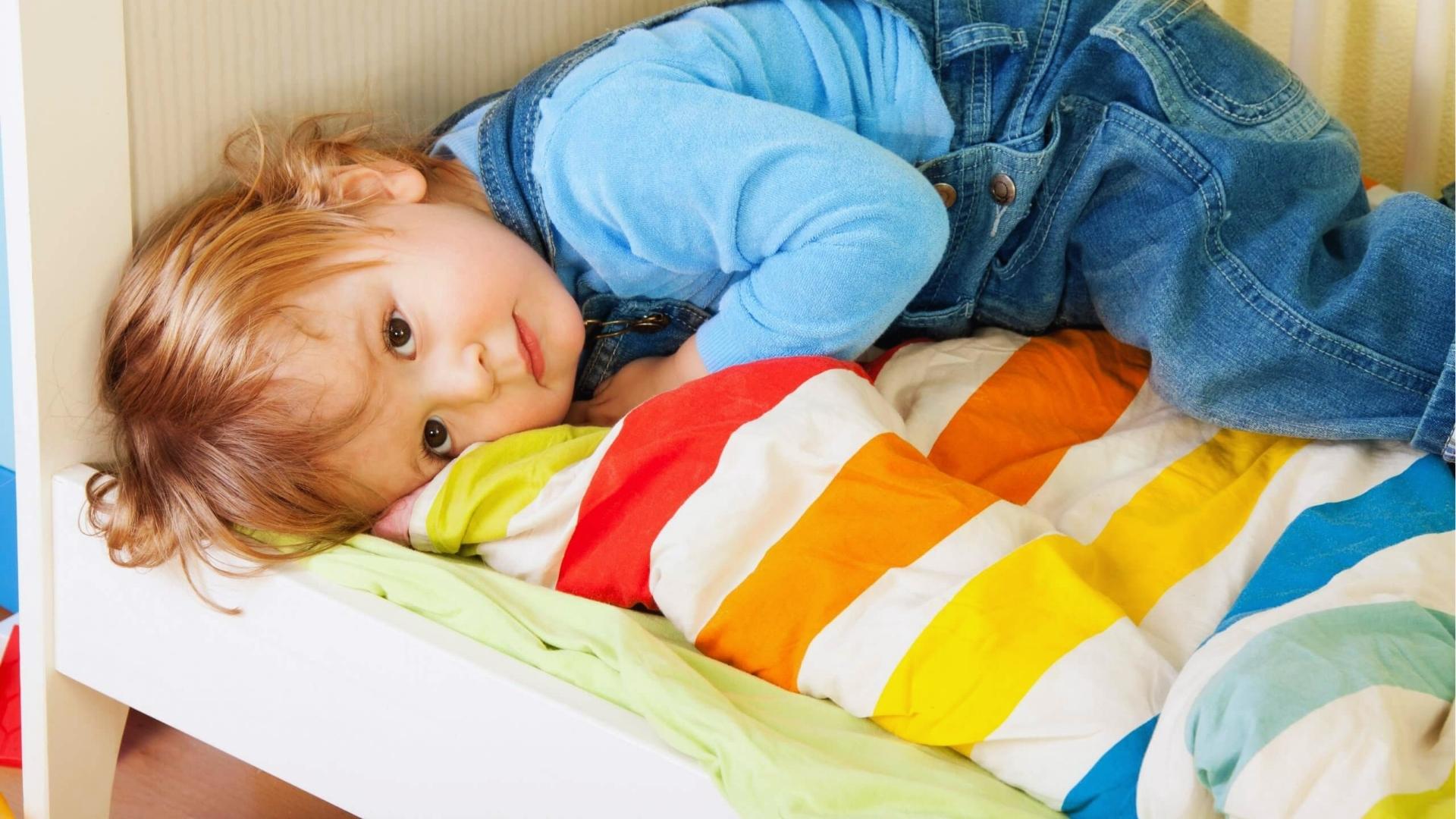 Toddler in blue overalls lies on bed with rainbow striped blanket, calm space, Activities for Autistic Kids.